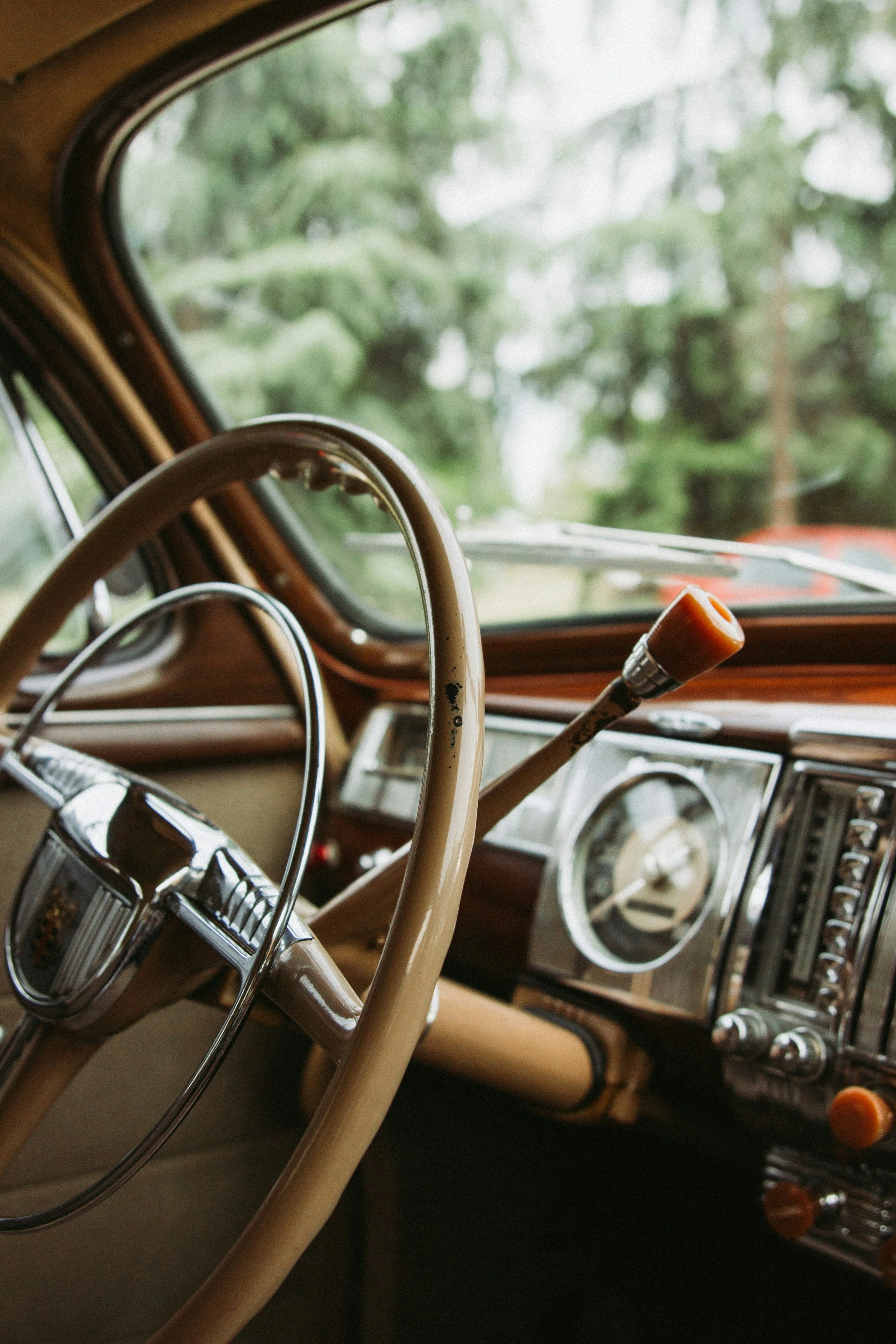Close-up view of the interior of a vintage car, showing a beige steering wheel, dashboard, and gear shift, with trees visible through the windshield.