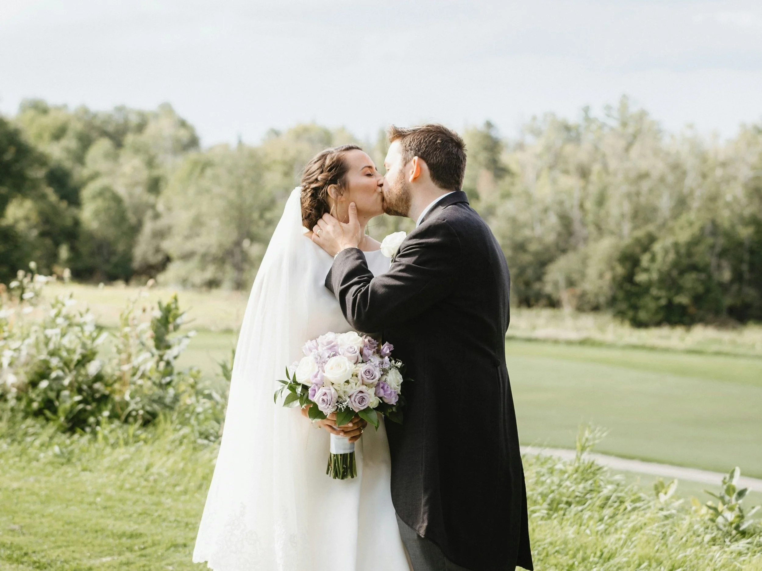 A bride and groom sharing a kiss outdoors on their wedding day, with greenery and trees in the background. The bride is holding a bouquet of white and lavender roses.