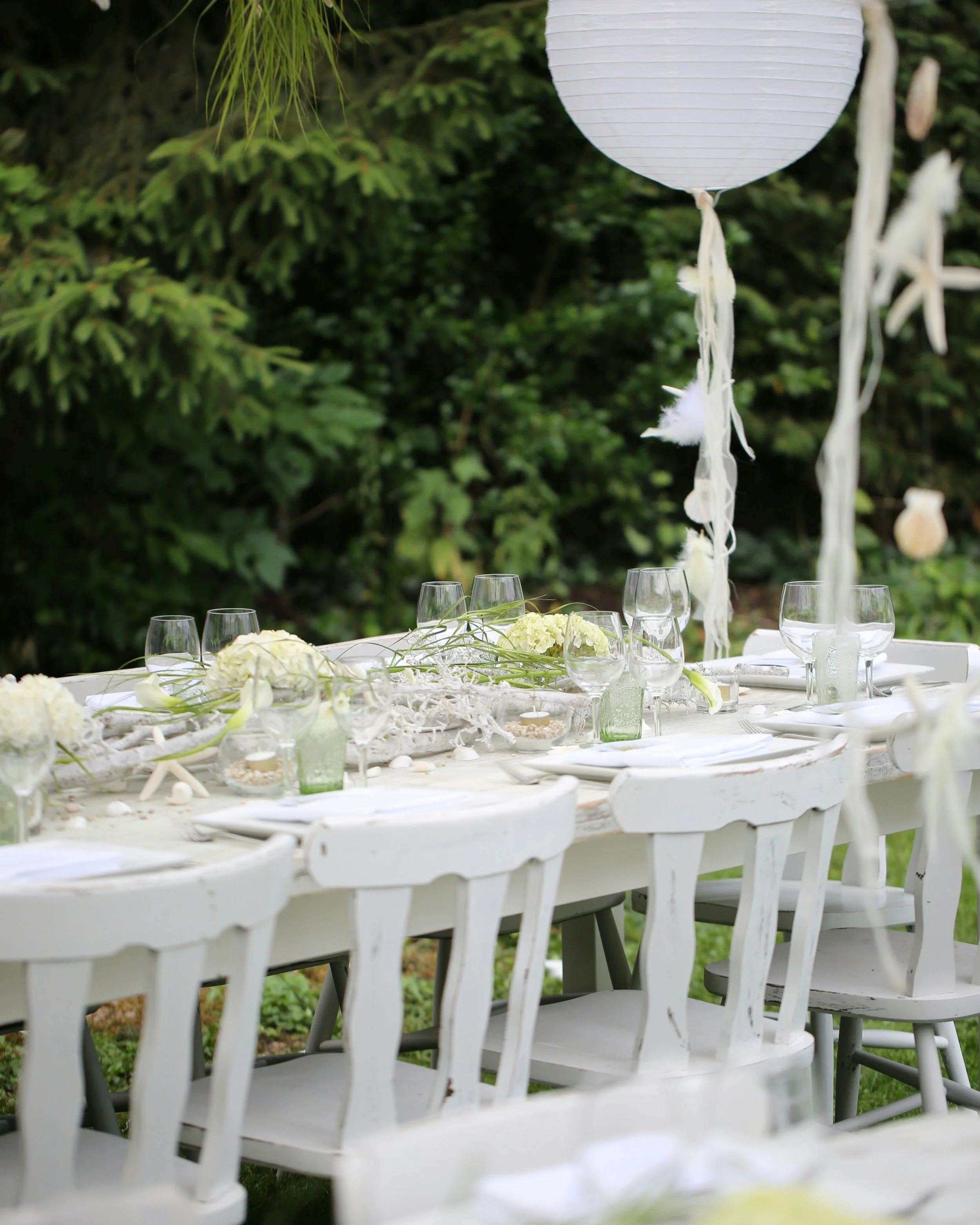 A decorated outdoor table with white chairs, flowers, and a hanging paper lantern in a garden setting.