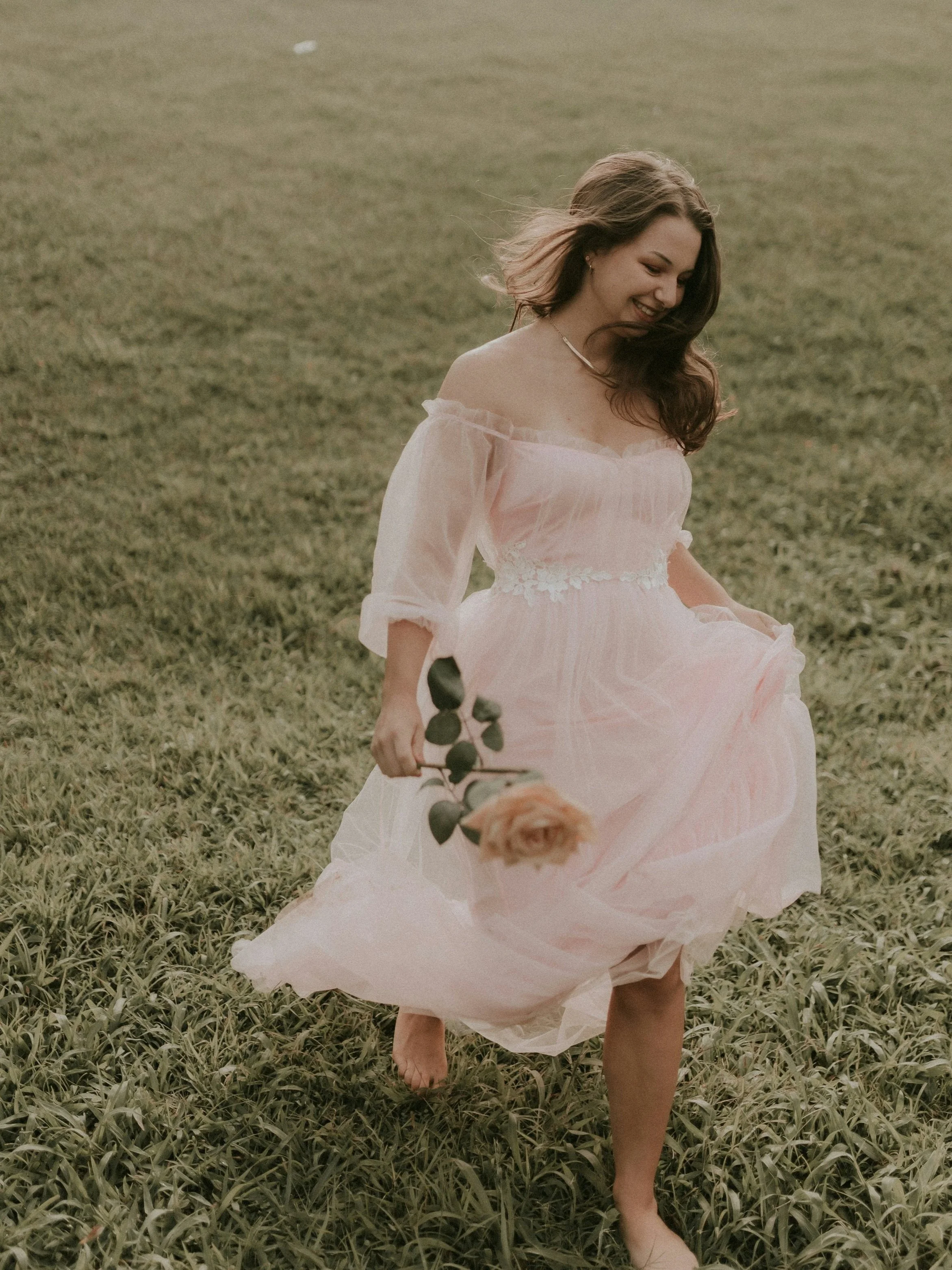 A woman in a light pink dress walking barefoot through a grassy field, holding a pink rose, with her hair flowing and a smile on her face.