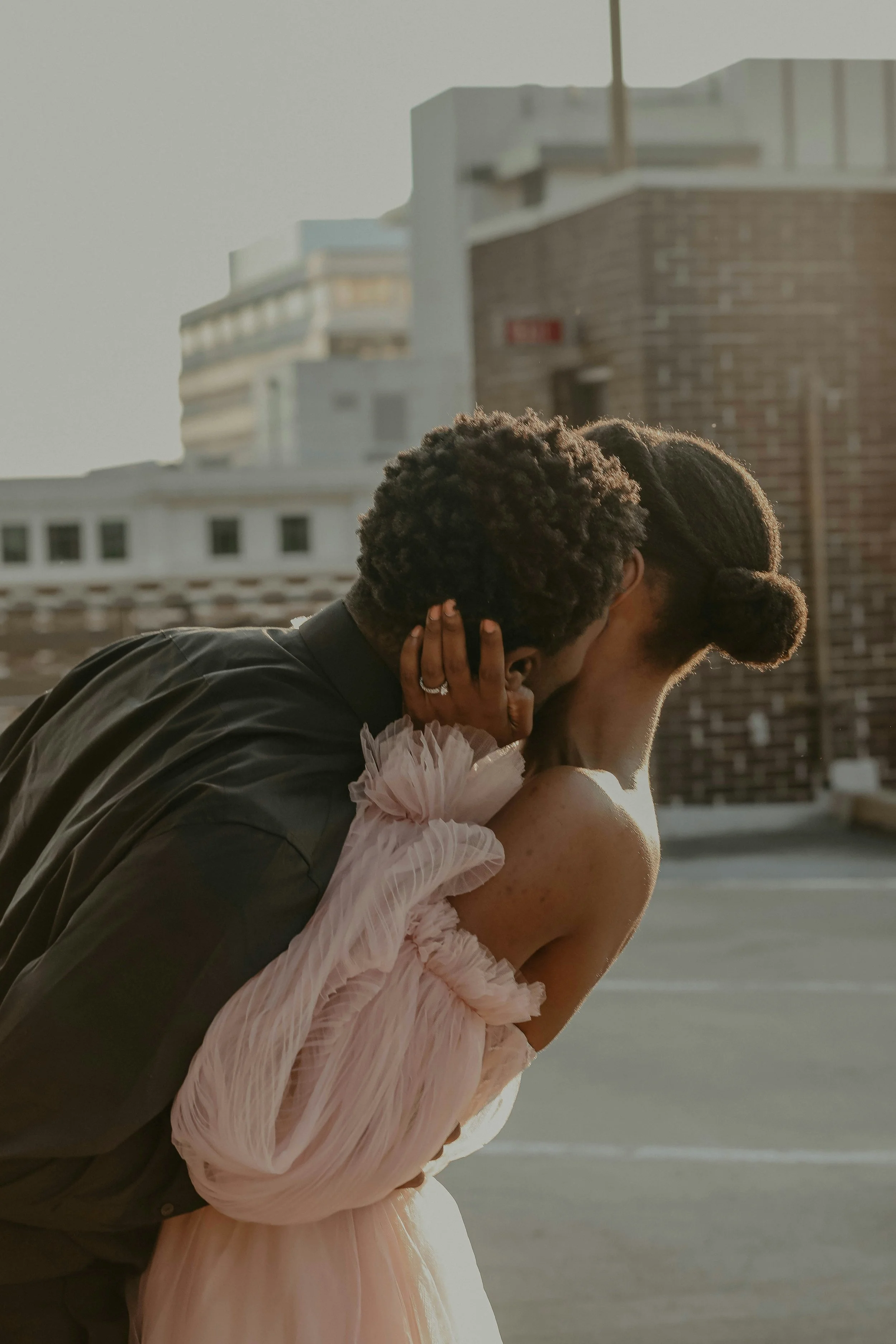 A man and woman sharing an intimate moment outdoors, with the man leaning over the woman and the woman holding his face with her hands, both partially wrapped in soft, pink, ruffled clothing, with a cityscape of buildings in the background.