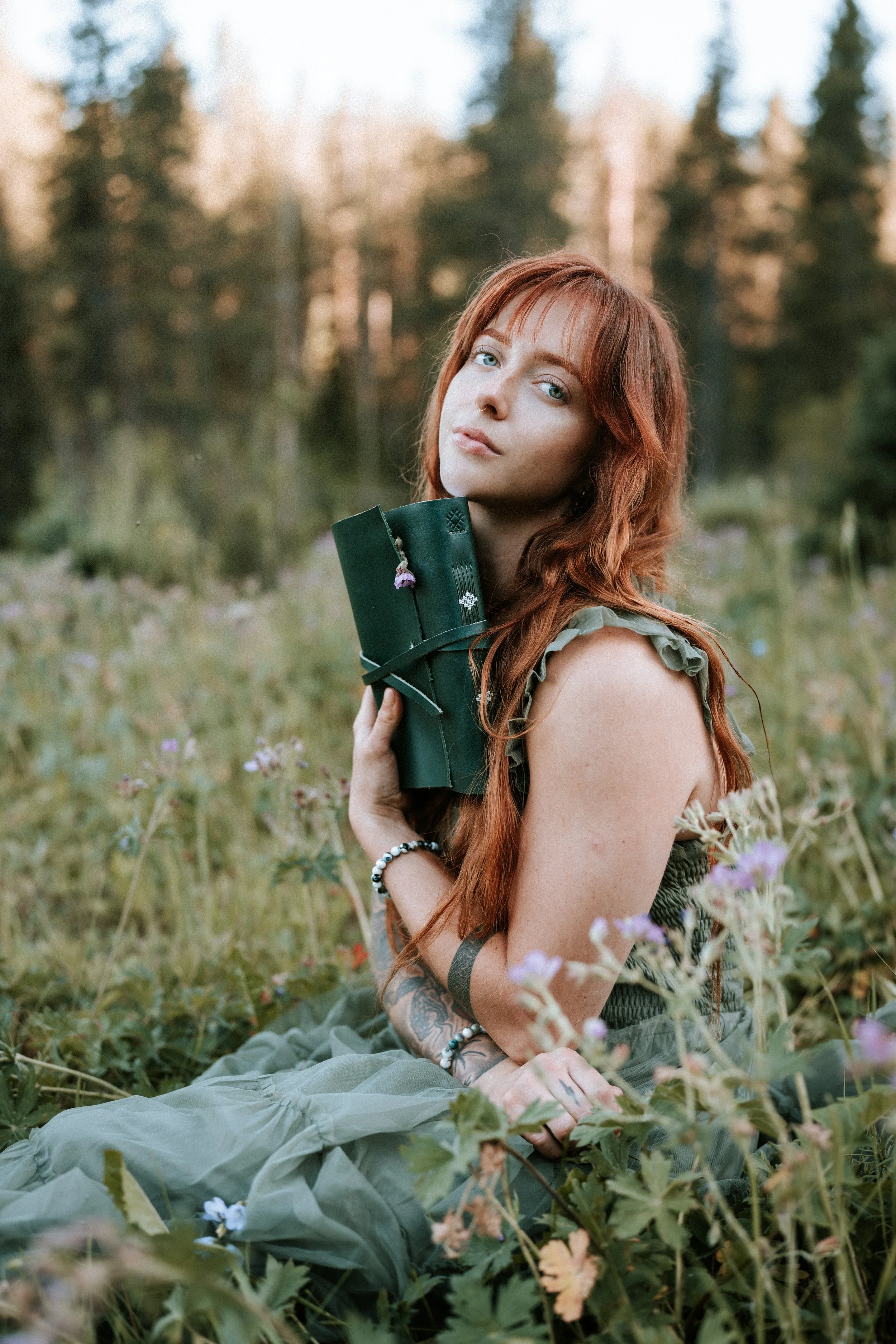 A young woman with red hair and tattoos sitting in a grassy field, holding a rolled journal or notebook, with trees in the background at sunset. Montana editorial portrait photographer.