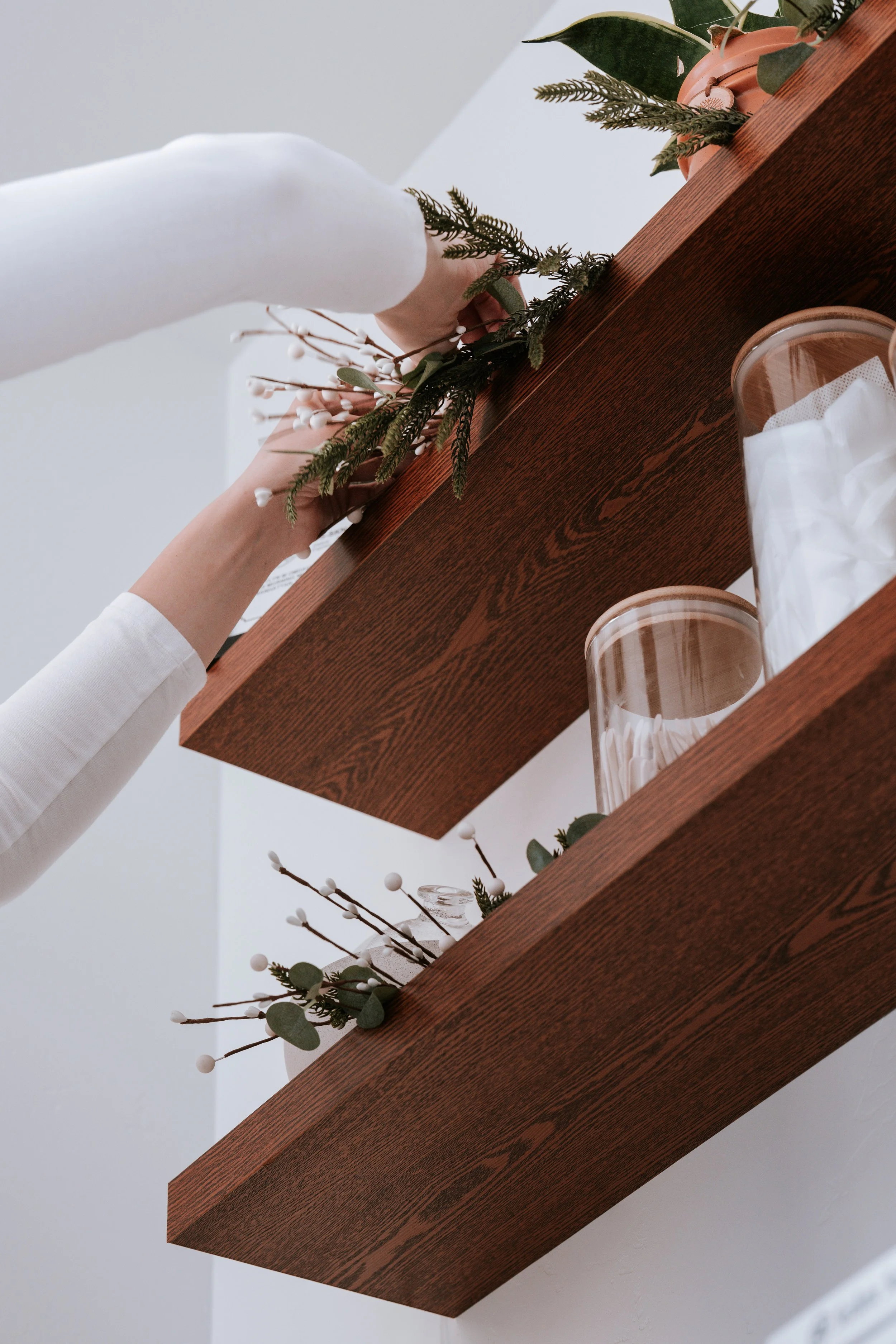 Person decorating wooden wall shelves with greenery and white accents, with glass jars on the shelves. Montana small business branding headshots photographer.