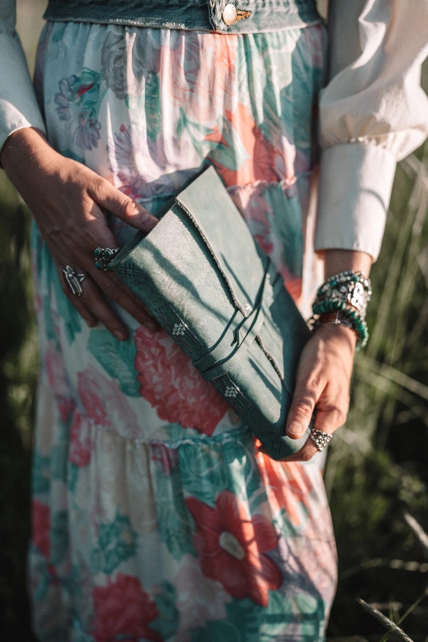 Person holding a small green clutch purse while wearing a floral skirt and white blouse, with jewelry on their hands and wrists. Montana editorial portrait photographer.