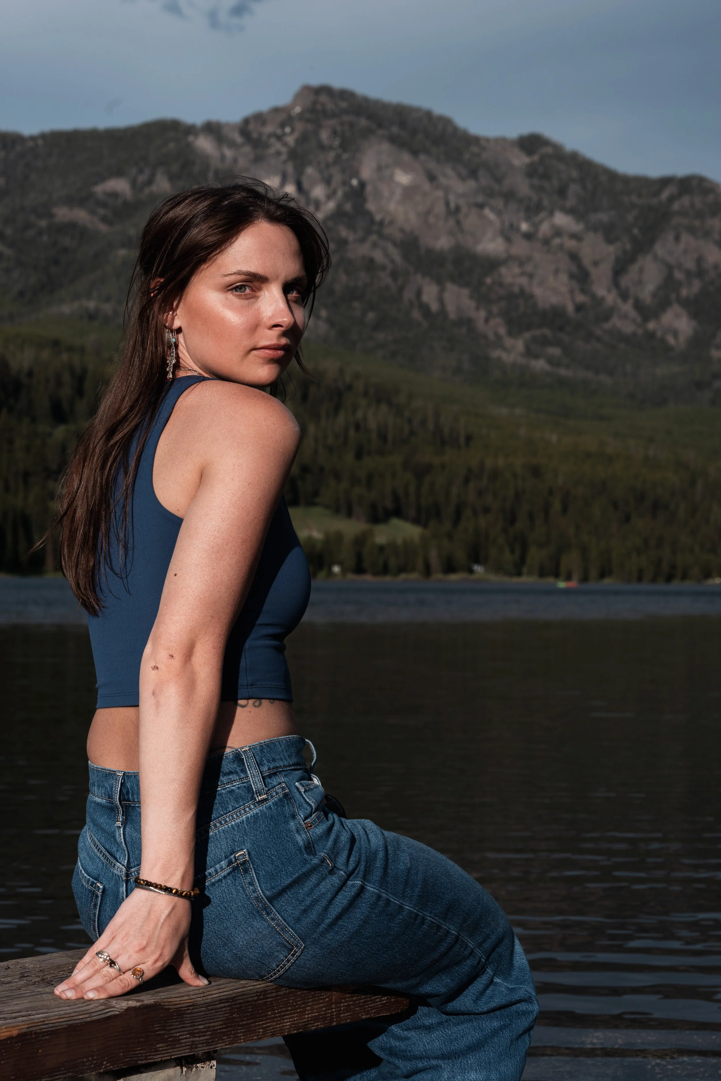 A young woman with long brown hair sitting on a wooden dock near a lake with mountains in the background. Montana editorial portrait photographer.
