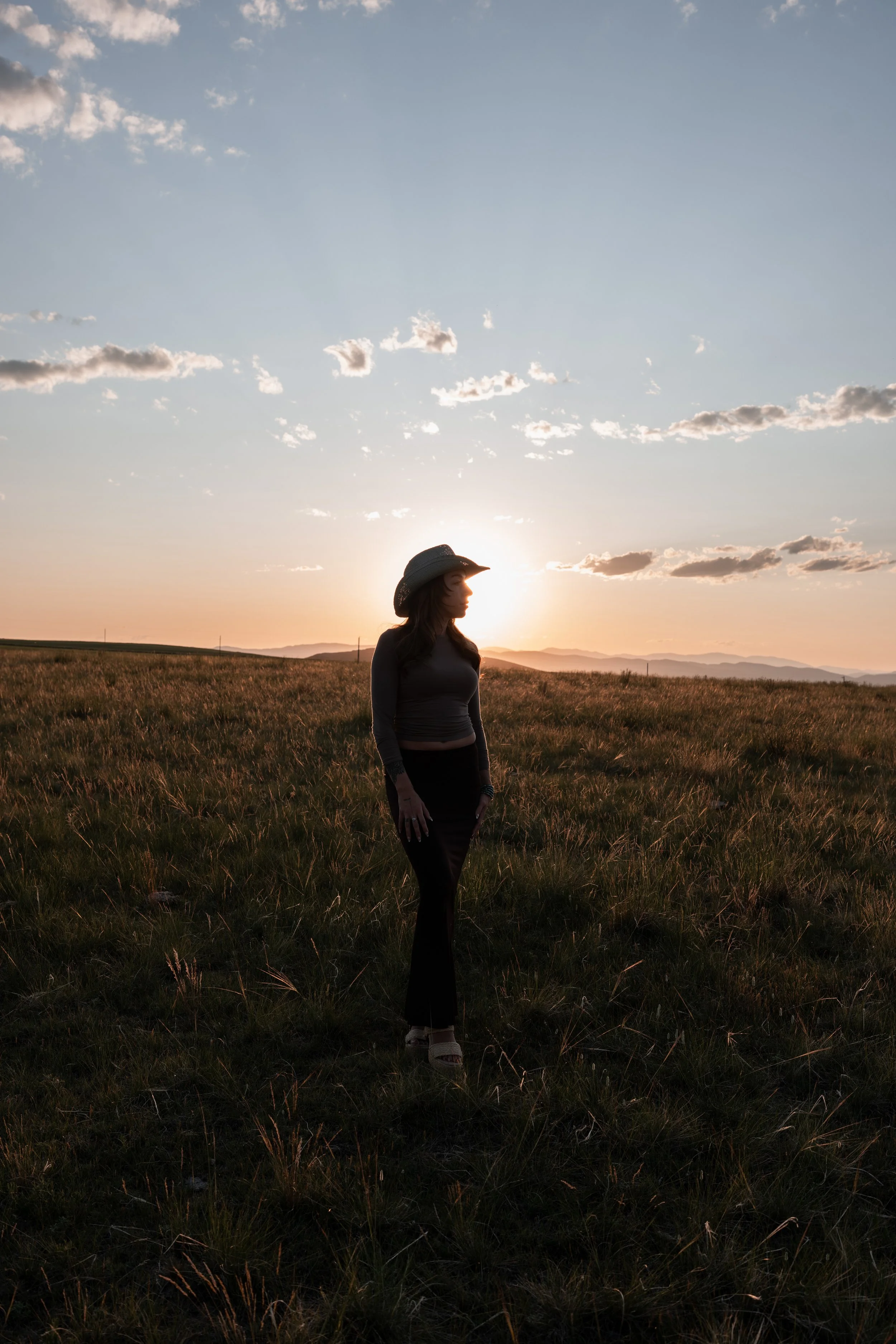 A woman wearing a wide-brimmed hat, standing in a grassy field during sunset. Montana editorial portrait photographer.