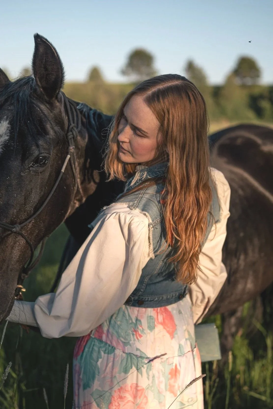A woman with long red hair, wearing a denim vest and a floral skirt, standing next to a black horse and gently holding its reins in an outdoor grassy field during daylight. Montana editorial portrait photographer.