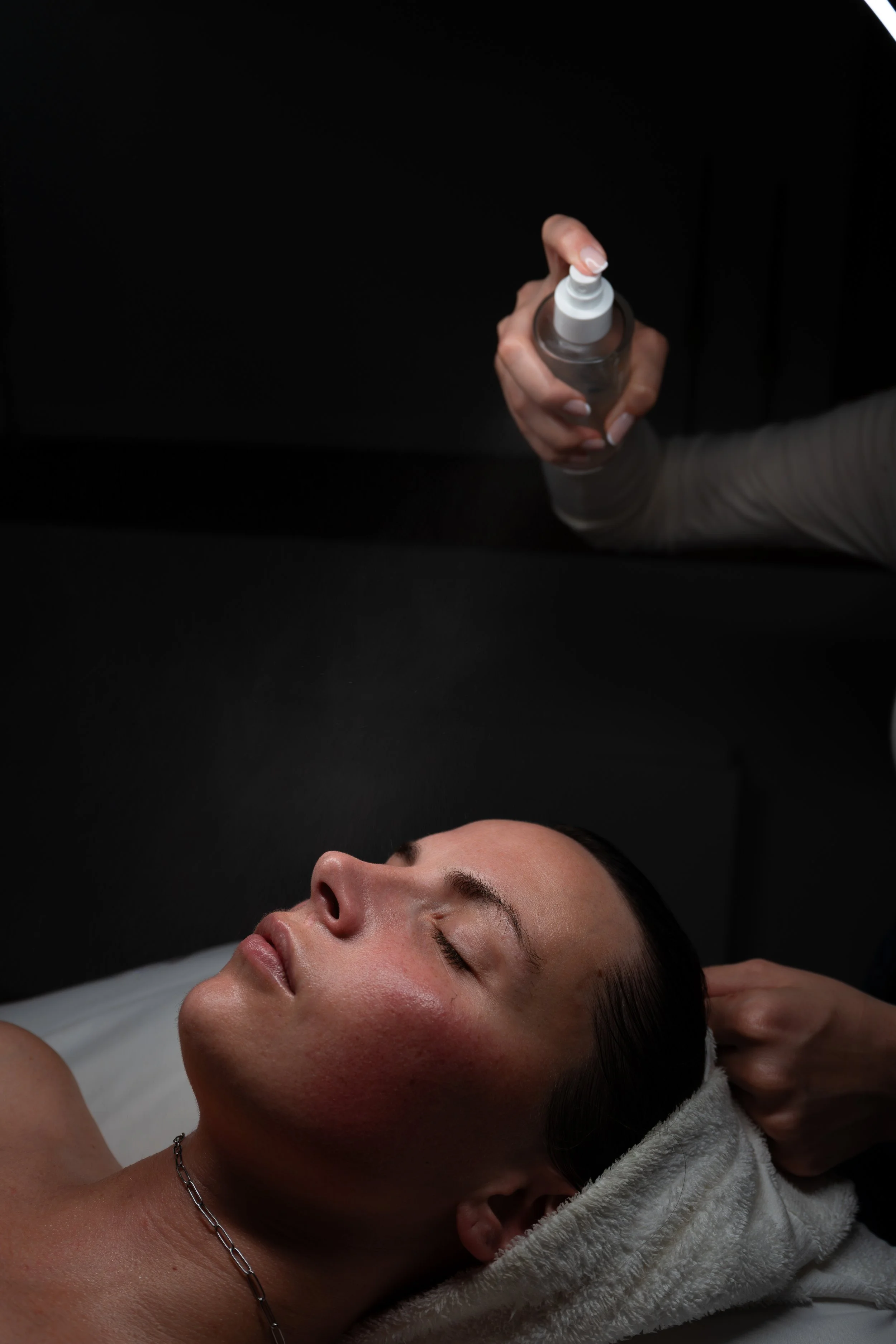 A woman lying face up with her eyes closed, receiving a facial treatment in a dark room, with a stylist or therapist holding a spray bottle above her face. Montana branding photographer for small business.