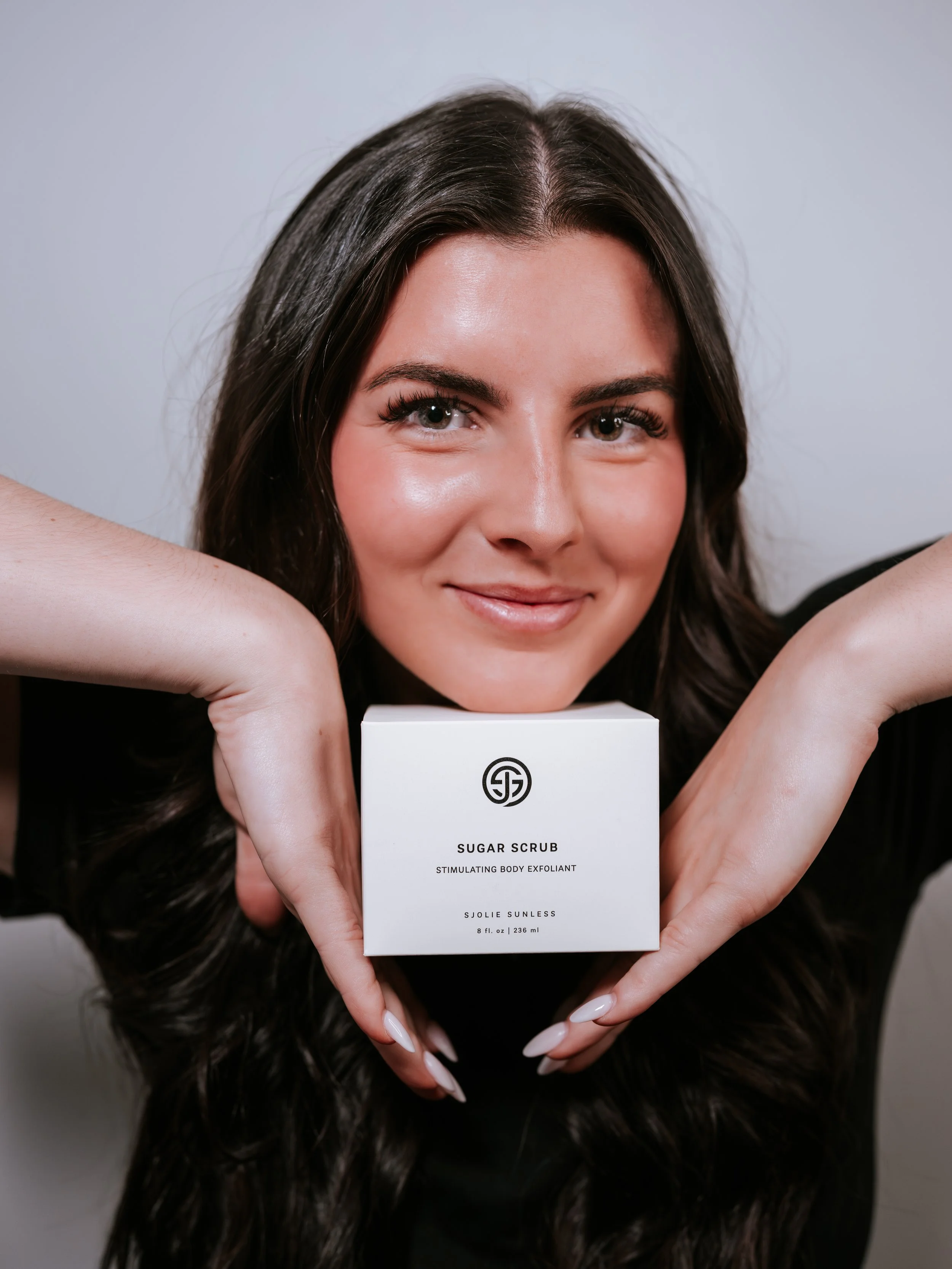 Esthetician with long, dark hair smiling while holding a box of sugar scrub in front of her face. Montana small business headshots branding photographer.