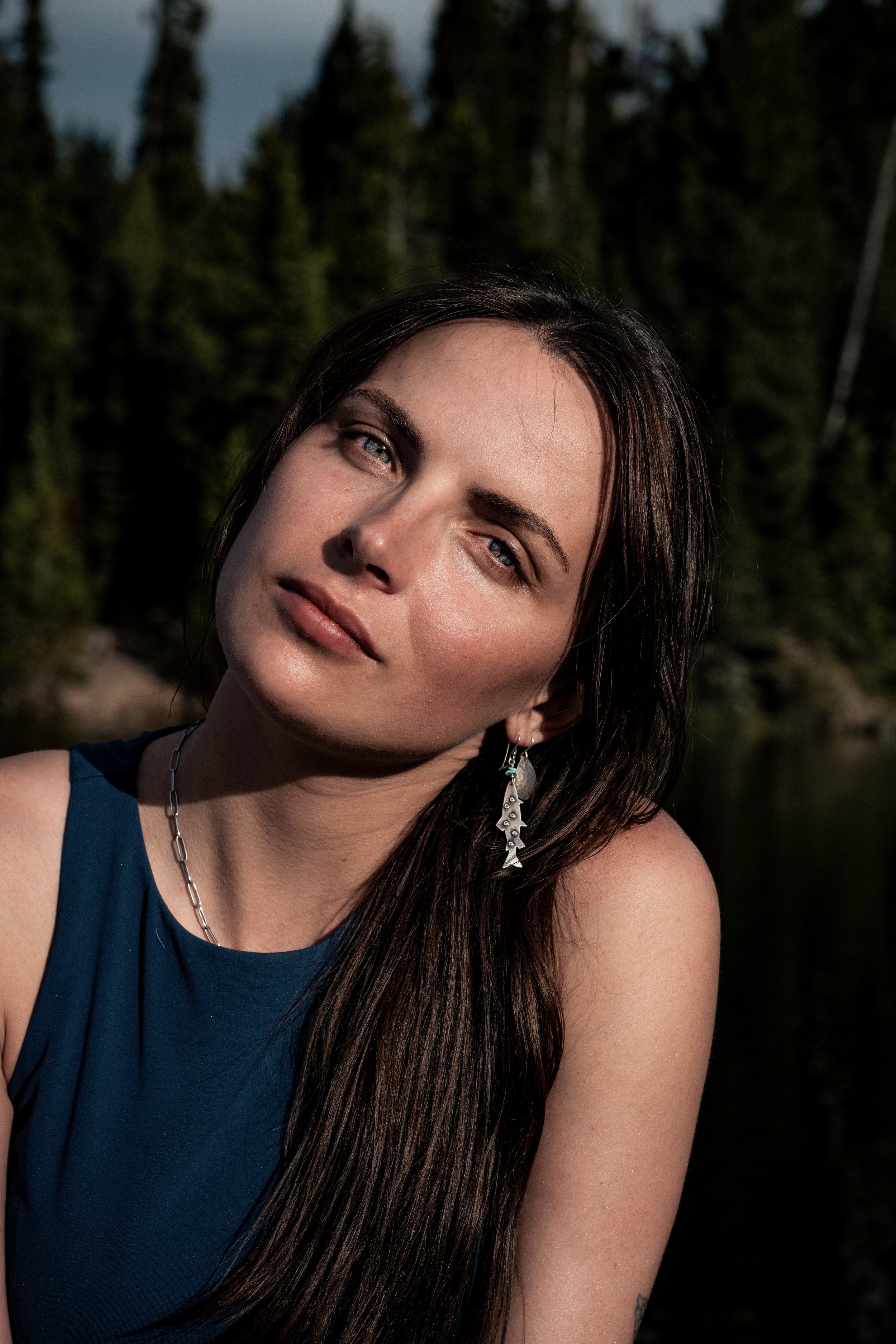 A woman with long dark hair, wearing a blue sleeveless top and earrings, tilts her head slightly to the right, outdoors with trees in the background. Montana editorial portrait photographer.