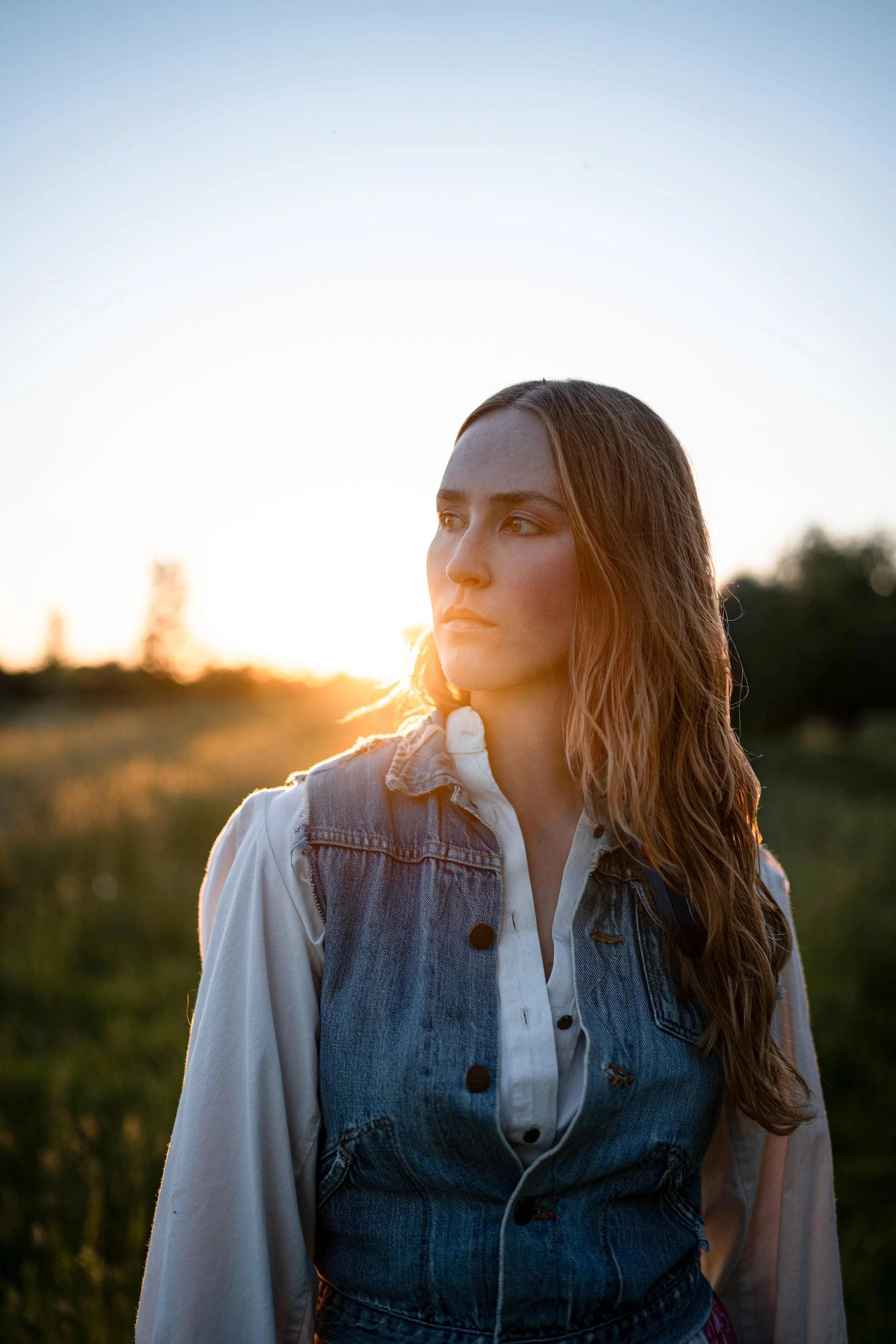 A woman with long, wavy hair standing outdoors during sunset, wearing a white shirt and denim vest, looking to the side. Montana editorial photographer.