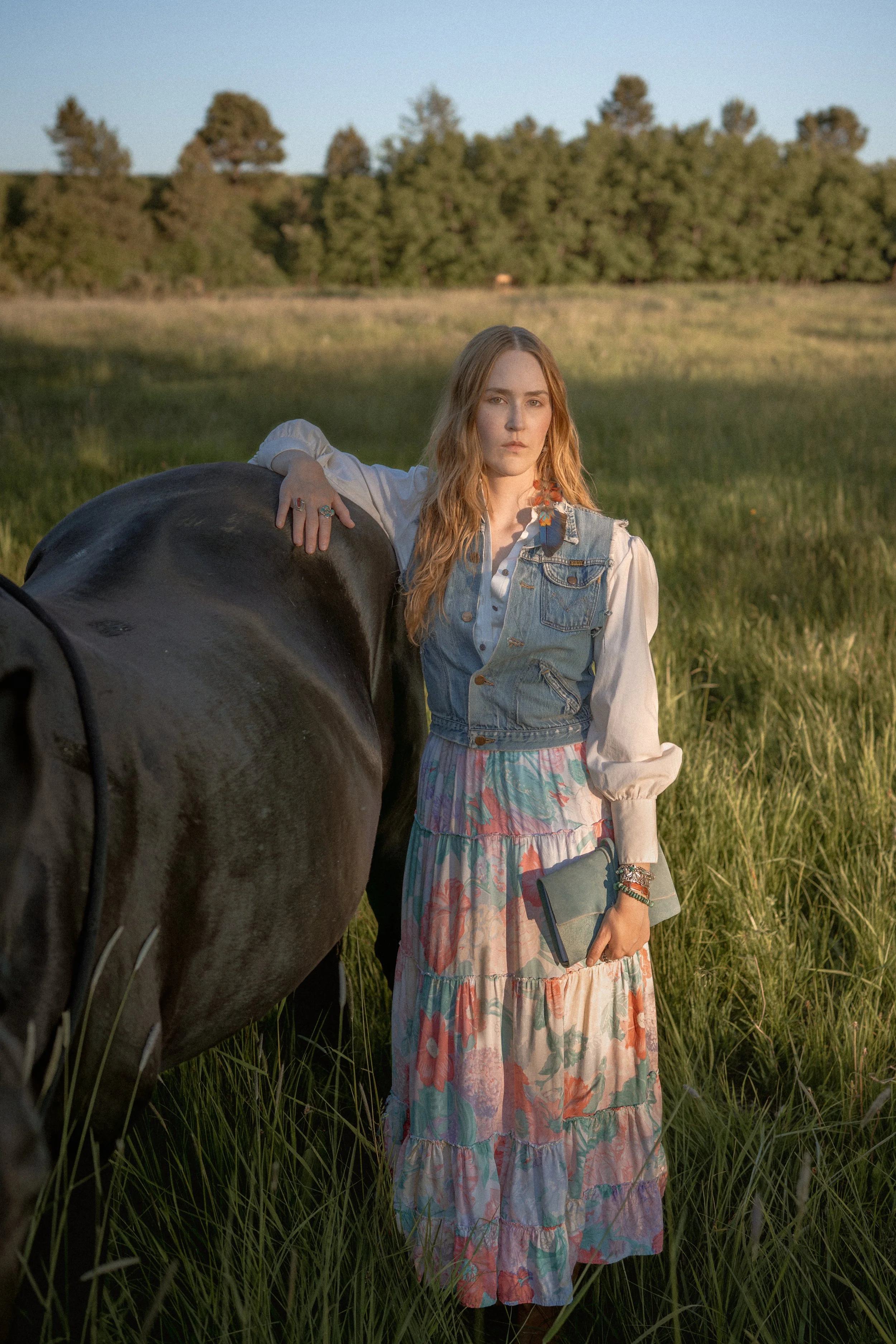 A woman with long red hair standing in a grassy field, resting her right arm on a black horse, holding a small clutch purse in her left hand, wearing a denim vest over a white blouse and a long, colorful floral skirt, with a scenic background of trees and a clear sky.