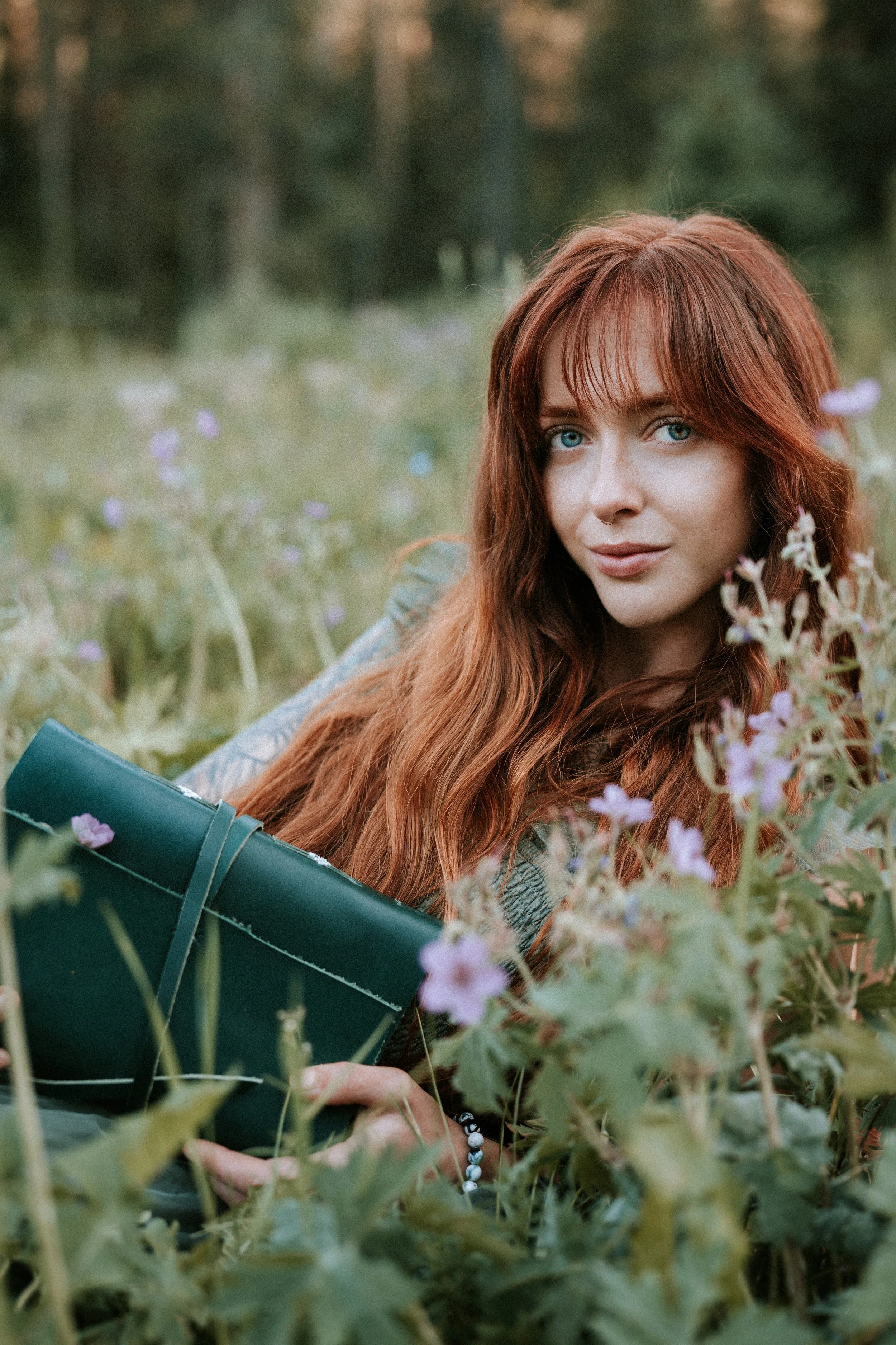 A woman with long red hair and blue eyes lying in a field of purple flowers, holding a closed green umbrella, with trees in the background.