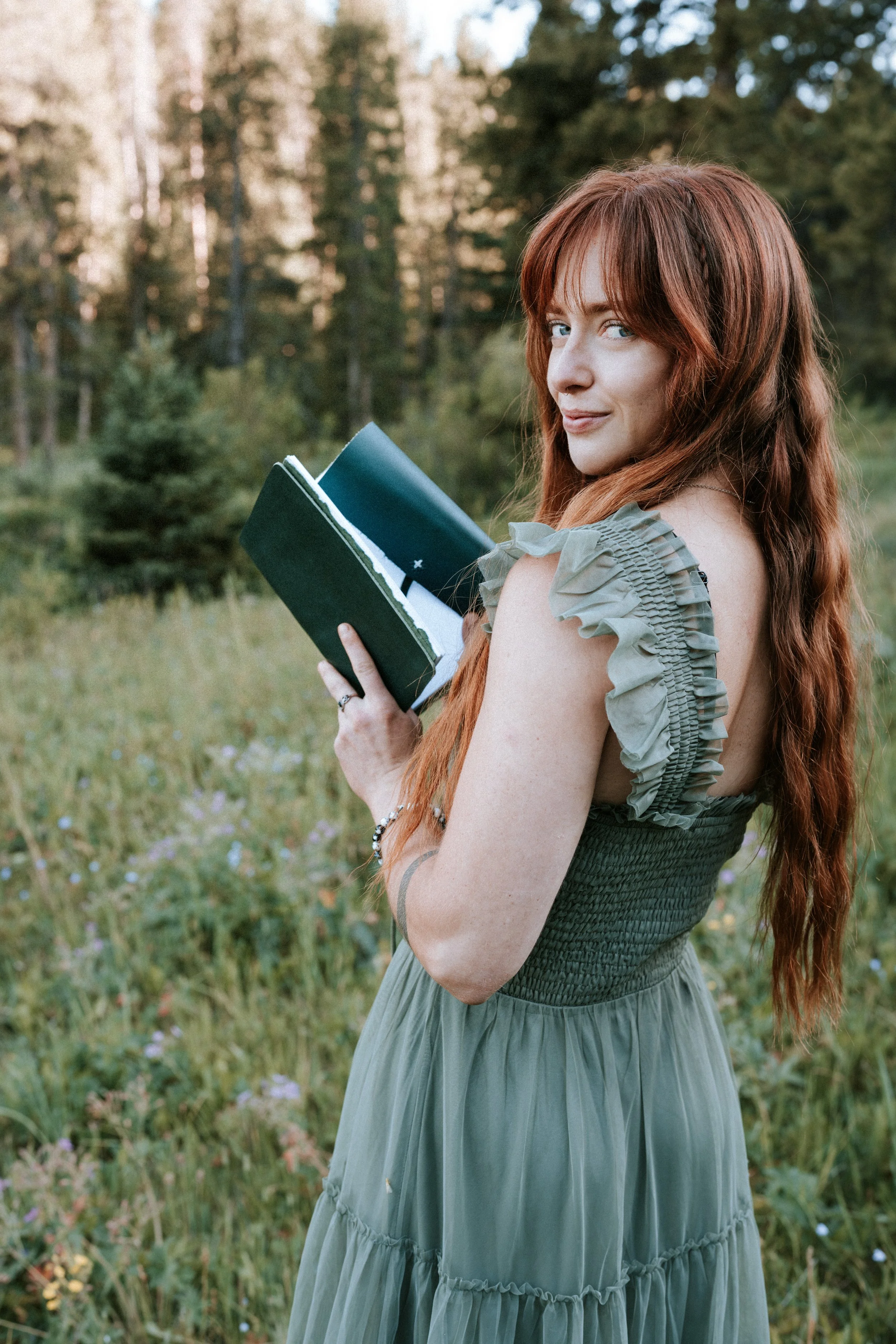 A young woman with long red hair, wearing a green dress with ruffled sleeves, stands outdoors in a grassy field with trees in the background, holding a closed notebook, looking at the camera. Montana editorial portrait photographer.