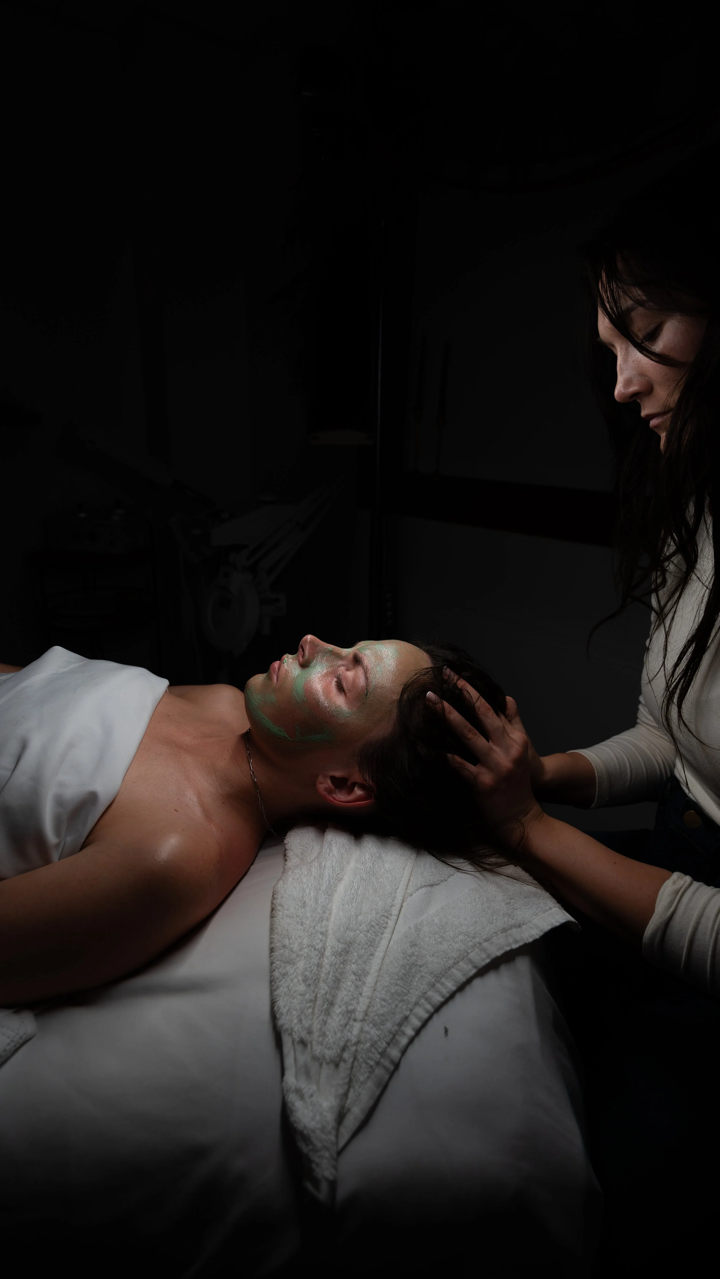 A woman lying on a massage table with her eyes closed, receiving a facial massage from a cosmetologist in a dimly lit room. Montana small business branding headshots photographer.