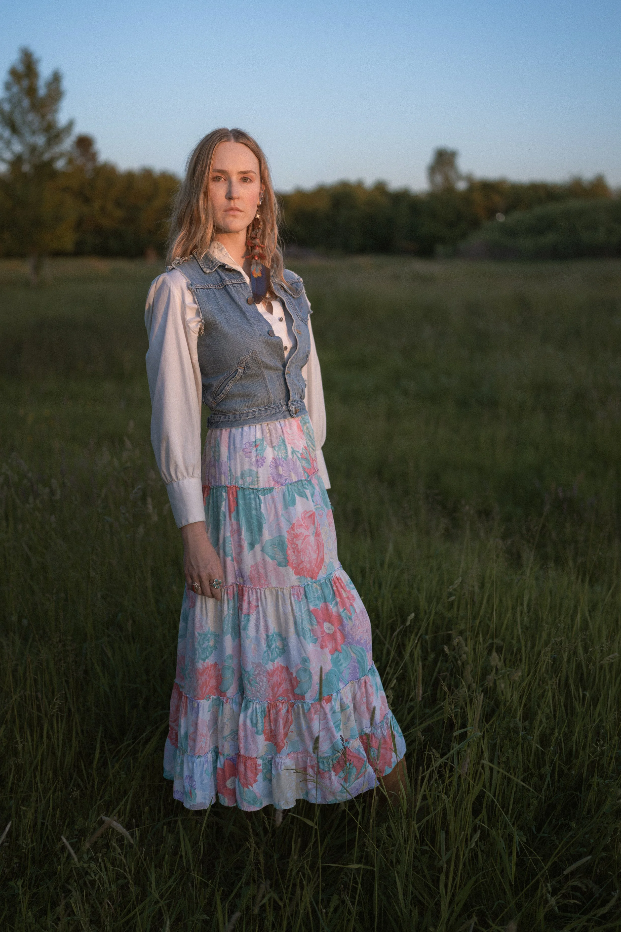 A woman standing in a grassy field at sunset, wearing a layered floral skirt, a white blouse, and a denim vest. Montana editorial photographer.