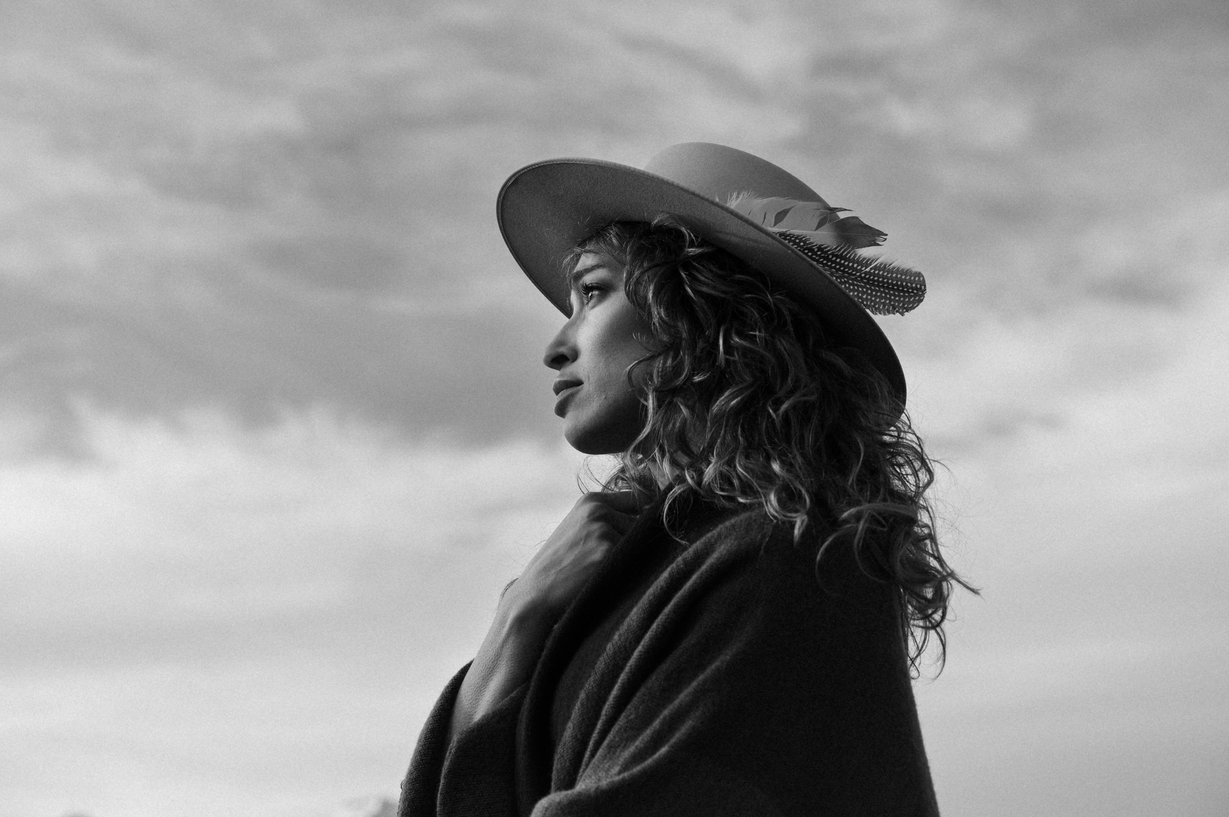A woman with curly hair wearing a wide-brimmed hat with feathers, standing outdoors with a cloudy sky in the background, looking to the left. Montana photographer.