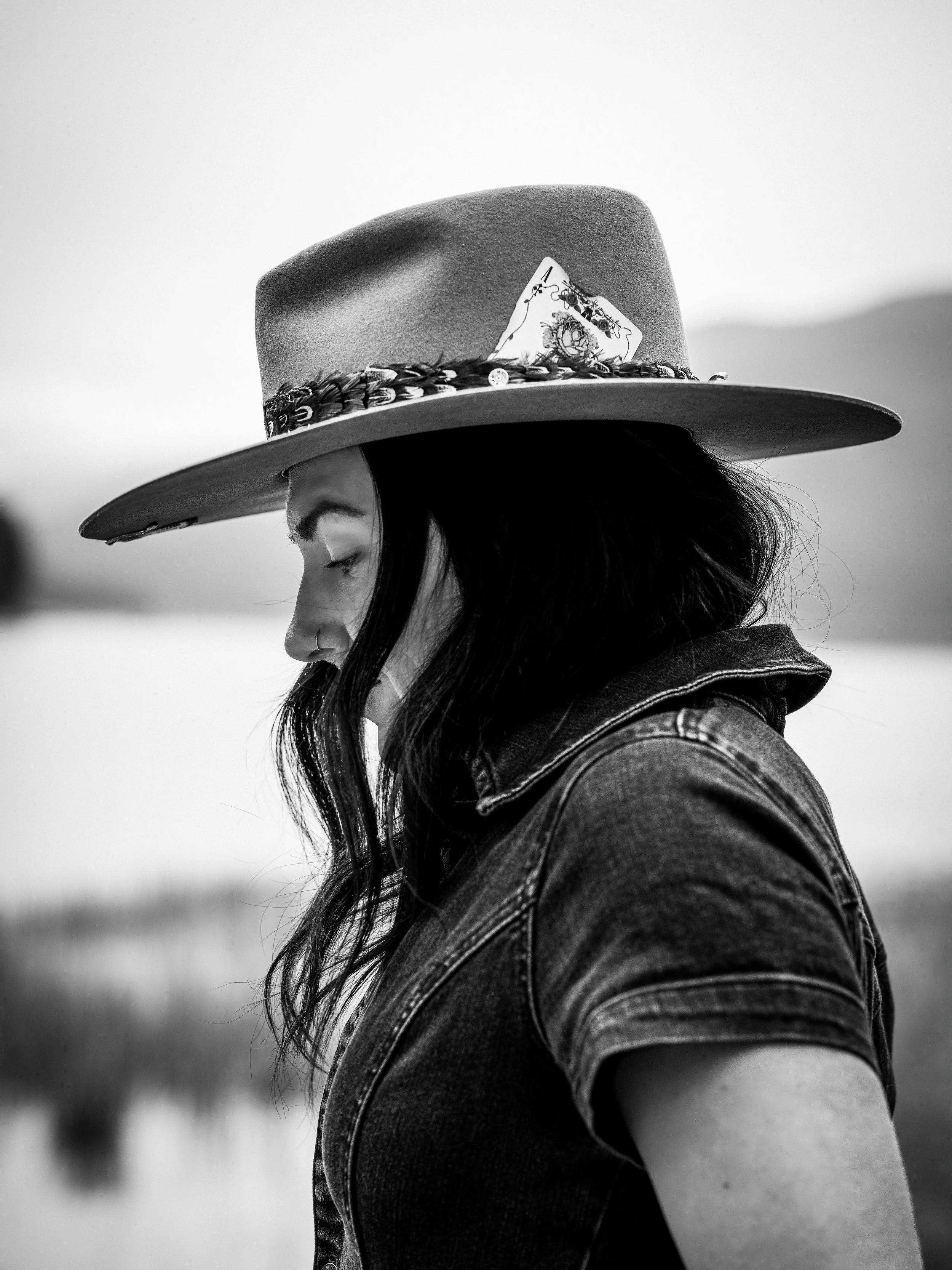 A woman with dark hair wearing a wide-brimmed hat decorated with a playing card, looking downward with her eyes closed, dressed in a denim jacket, outdoors near water. Montana editorial portrait photographer.