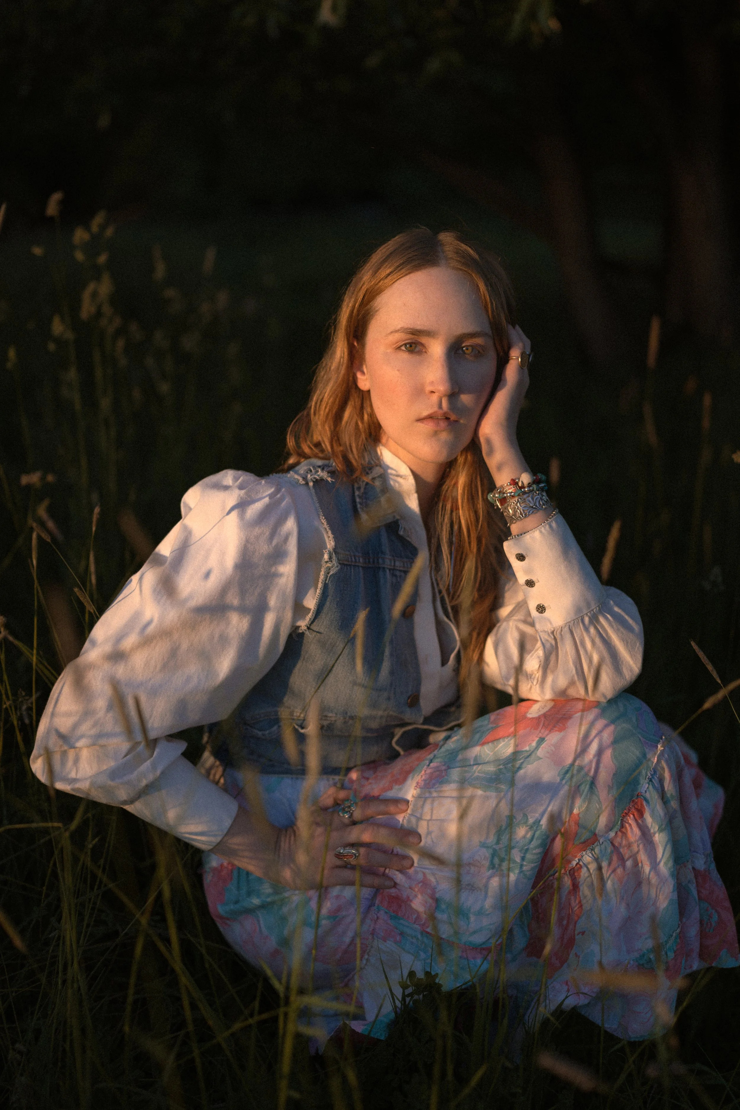 A woman with red hair and freckles sitting in tall grass during sunset, wearing a denim vest over a cream blouse and a floral skirt, with jewelry on her wrist and fingers, and looking thoughtfully at the camera.