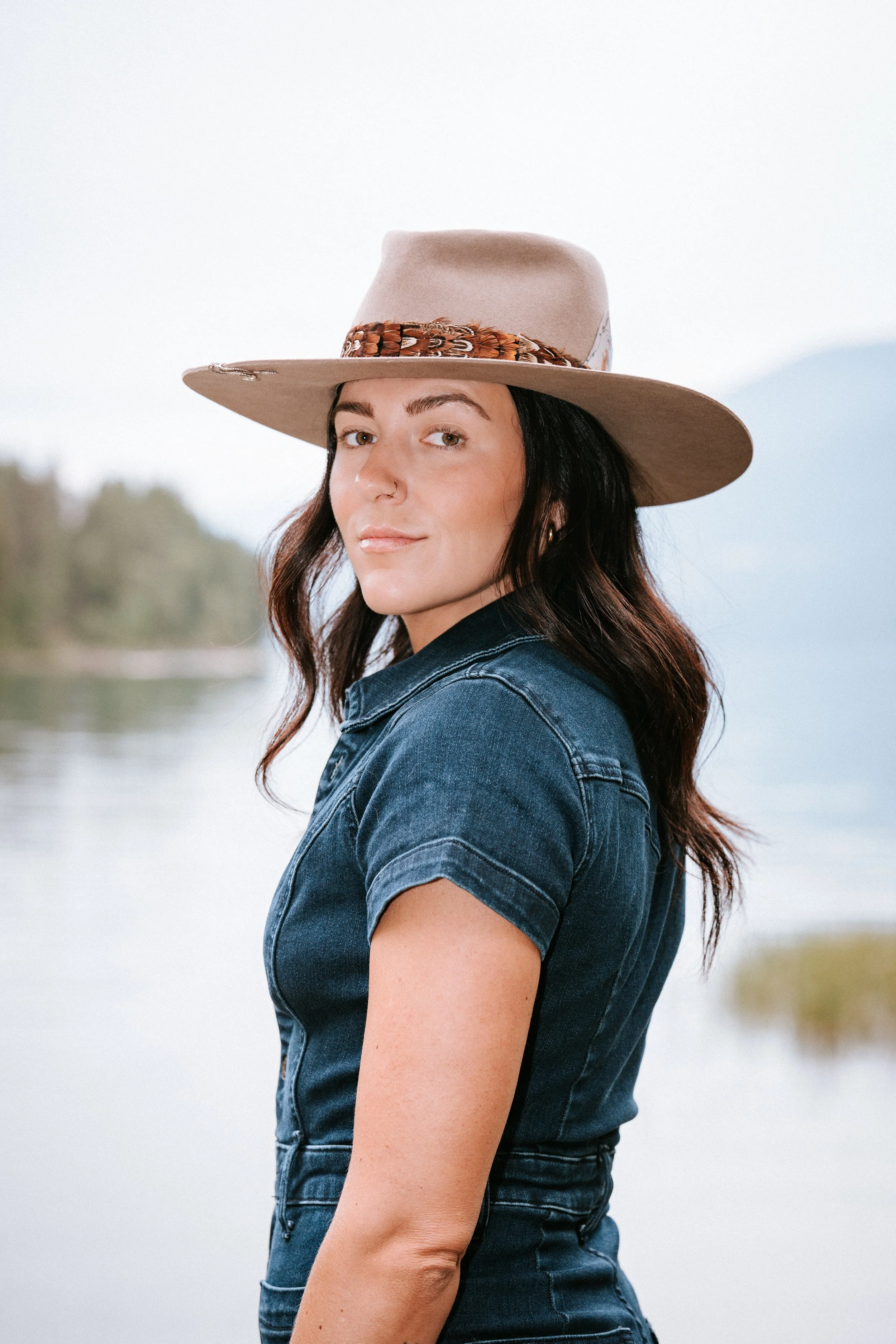 A woman with dark, wavy hair wearing a wide-brimmed beige hat with a decorative band and a denim shirt, standing outdoors near water with trees and mountains in the background. Montana portrait photographer.