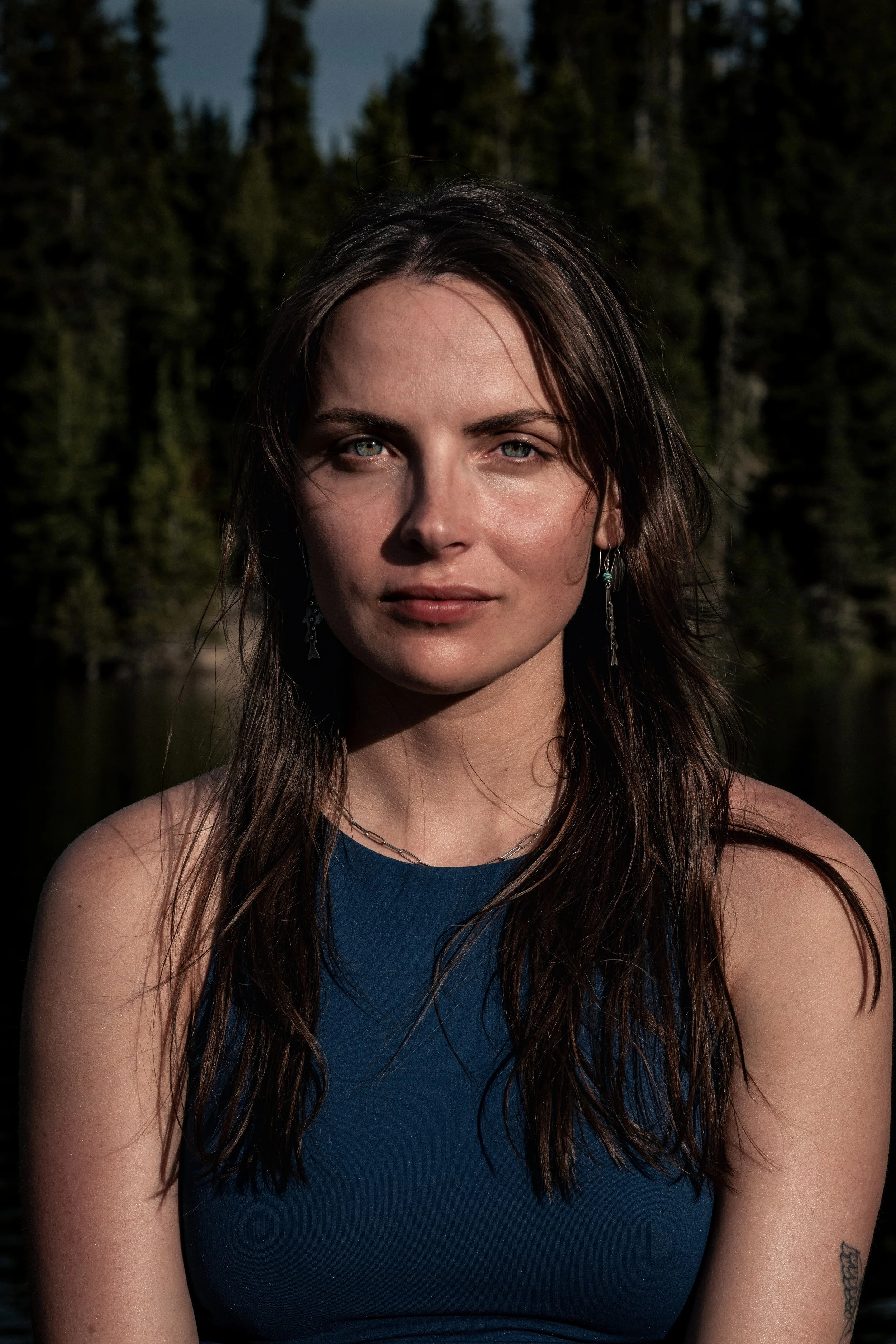 Close-up of a young woman with long brown hair, blue eyes, and earrings, outdoors with a forest background during daylight. Montana editorial portrait photographer.