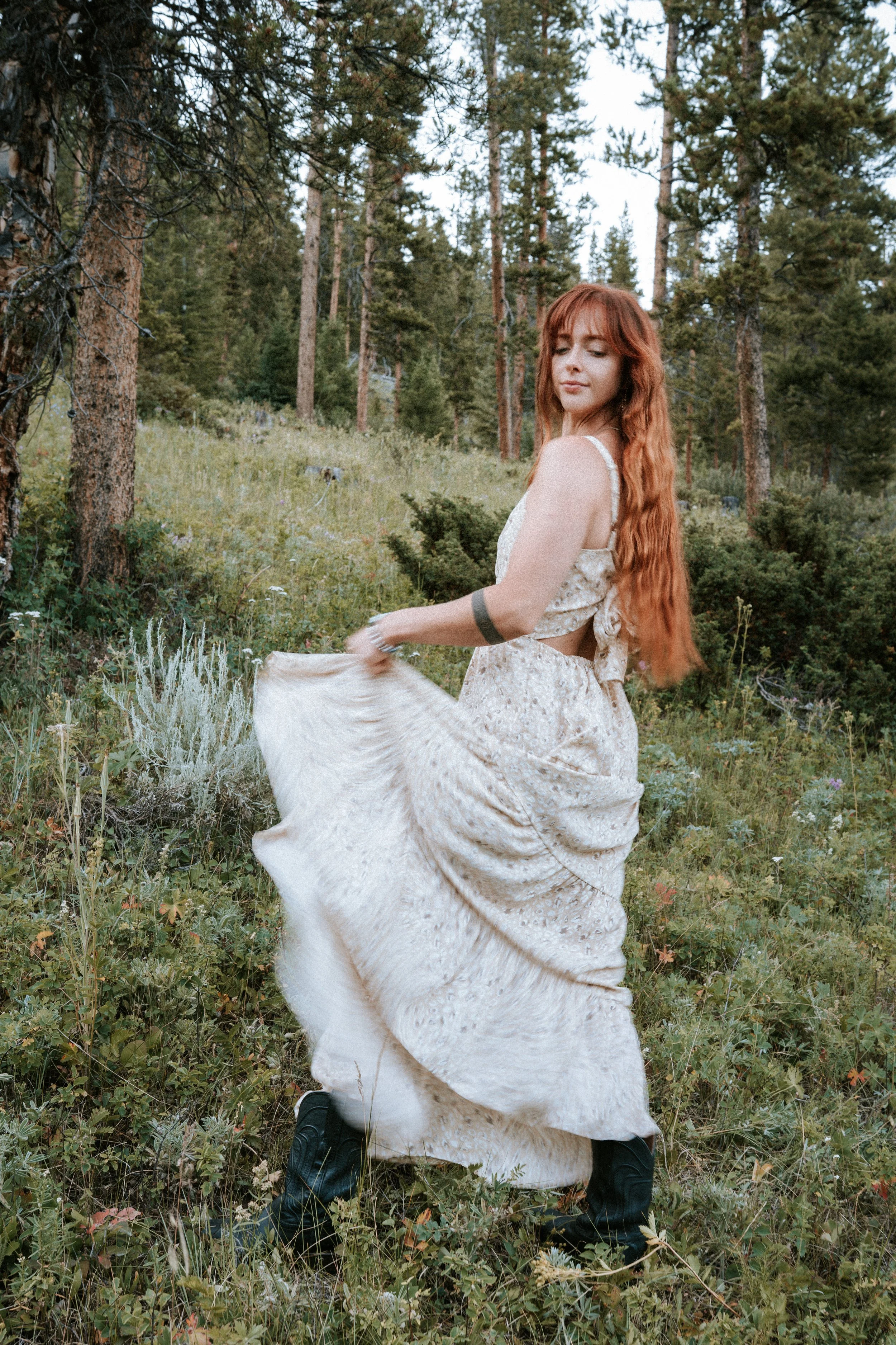 A young woman with long red hair wearing a light-colored dress and black boots, standing in a grassy forested area, holding her dress while looking at the camera with a calm expression. Montana editorial portrait photographer.