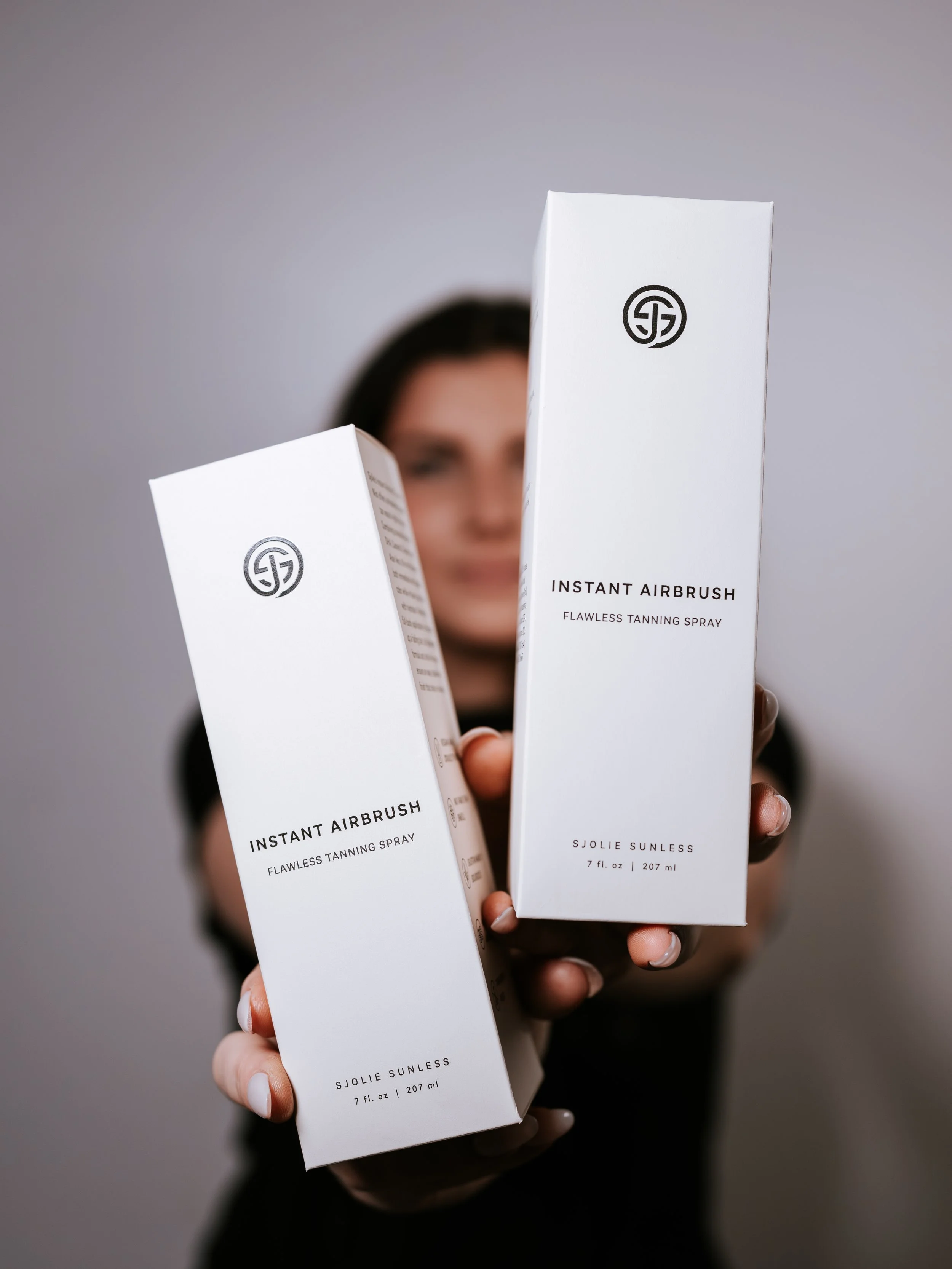 Esthetician holding two white boxes of S JOLIE SUNLESS instant airbrush self-tanning spray, with blurred background. Montana small business headshots branding photographer