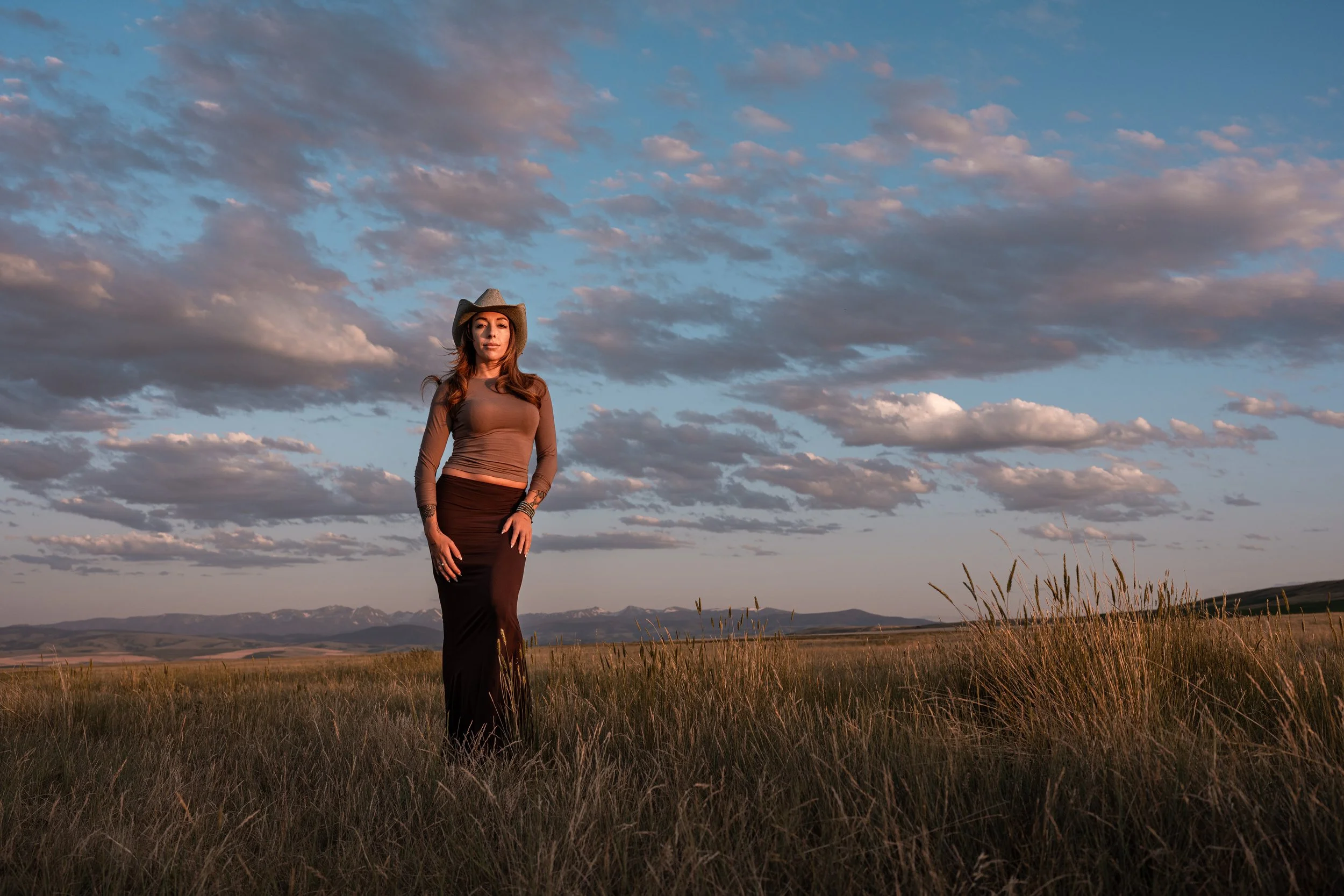 A woman wearing a cowboy hat and long skirt standing in open grassland at sunset, with a sky filled with clouds and distant mountains. Montana editorial portrait photographer.