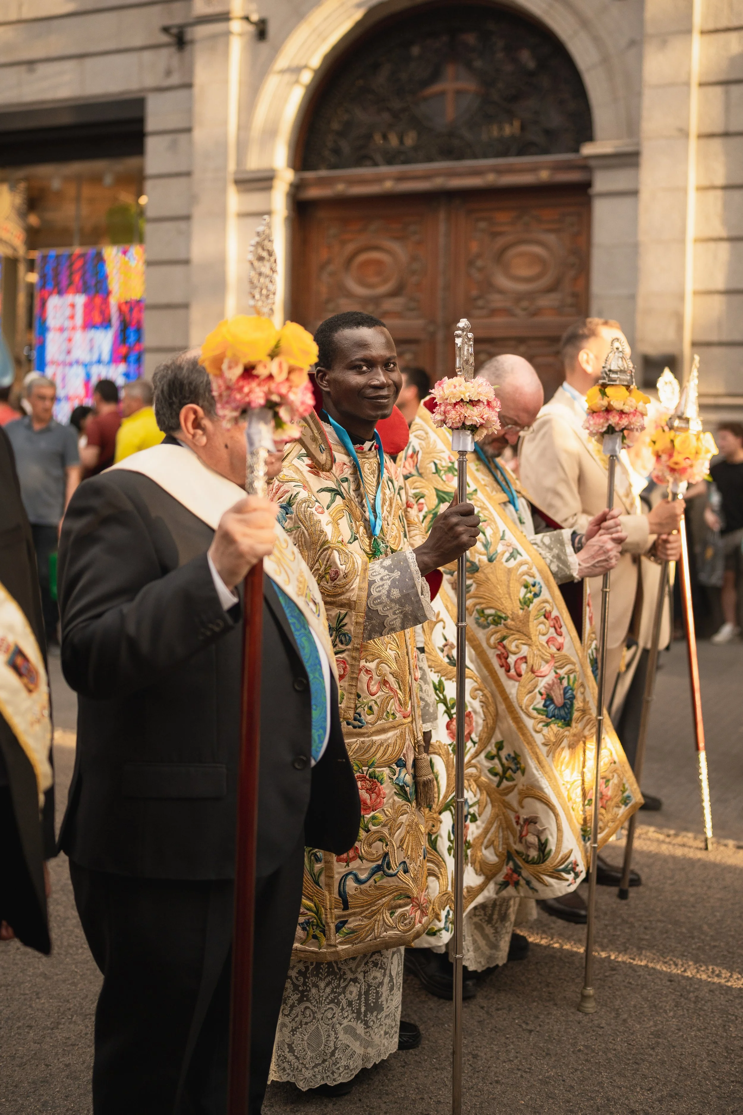 Group of men in ornate robes and suits holding decorated staffs during a religious procession in front of a church with an arched door. Montana editorial street photographer.