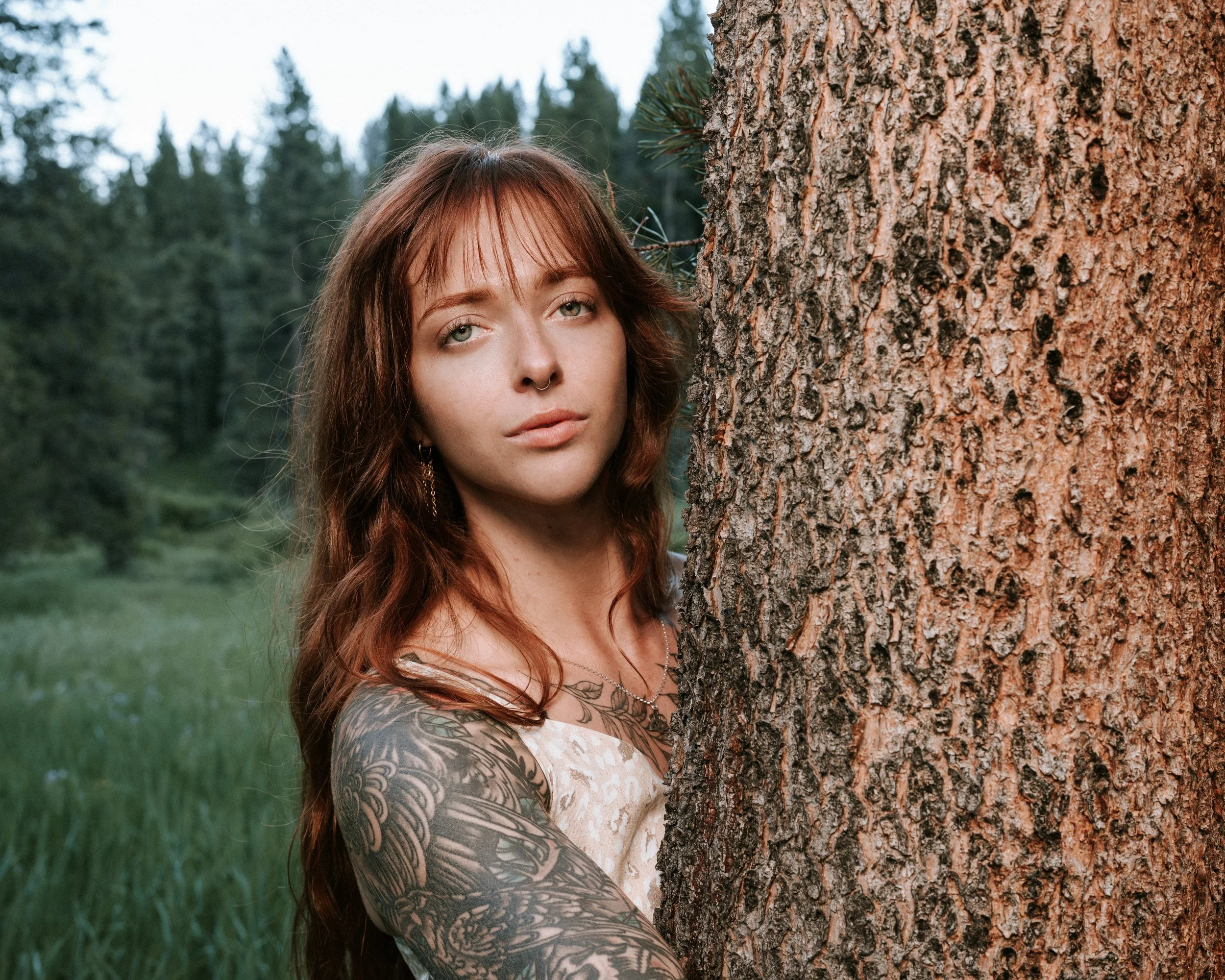 A young woman with red hair and tattoos on her arm stands behind a large tree in a forested area, looking at the camera.