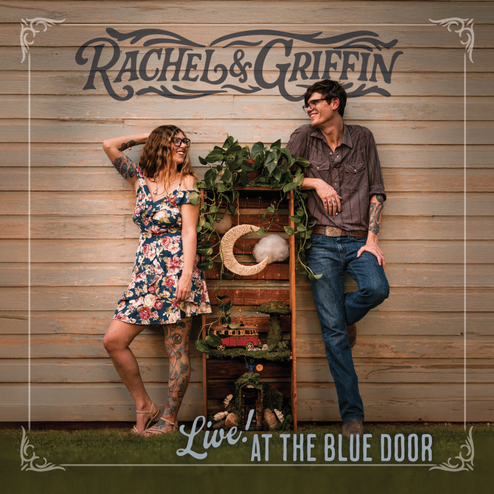 Album cover for “Live! at The Blue Door” by Oklahoma folk duo Rachel & Griffin, showing the musicians smiling beside a rustic wooden crate filled with miniature scenes and plants