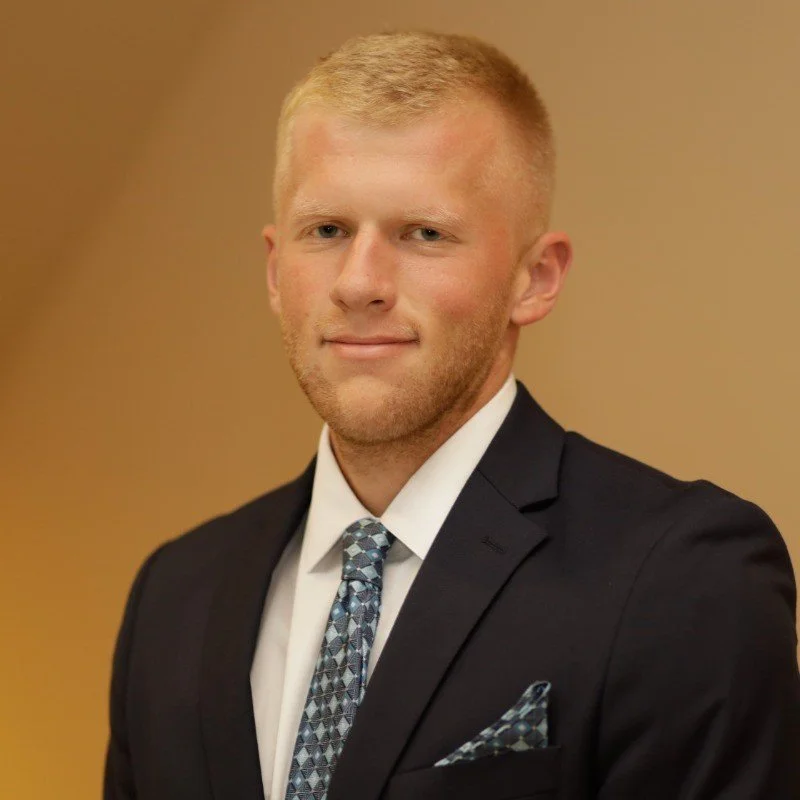 A young man with blond hair, light skin, and a slight smile, dressed in a formal black suit with a white shirt and a patterned blue tie and pocket square, against a neutral background.