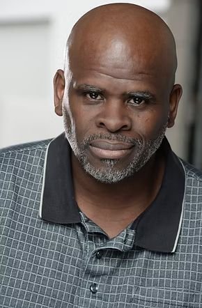 Close-up of a middle-aged African American man with a bald head and gray beard, wearing a dark collared shirt, with a serious expression in an indoor setting.