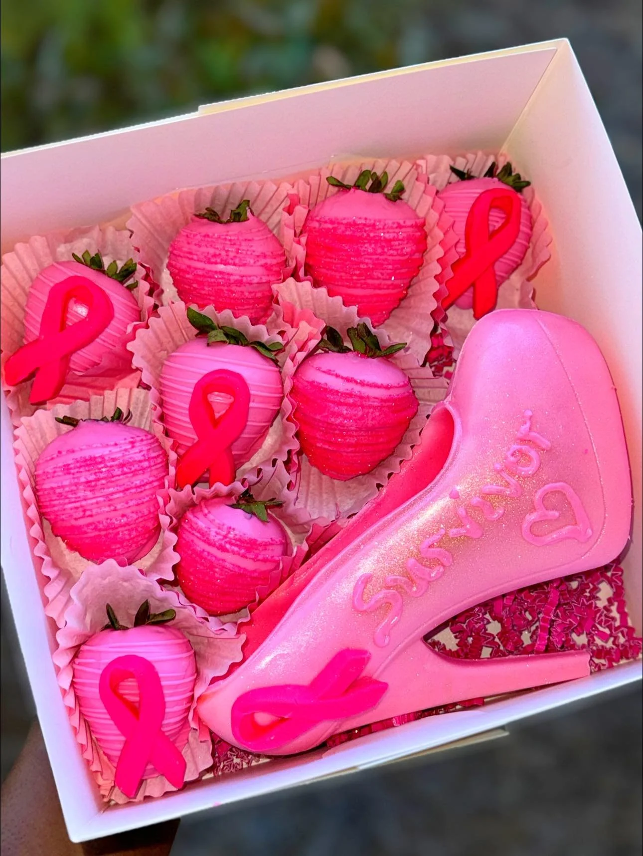 A white box containing pink strawberries decorated with pink ribbons, and a pink high-heeled shoe shaped cookie with the words 'Save the' written on it, along with a heart symbol, all themed for breast cancer awareness.