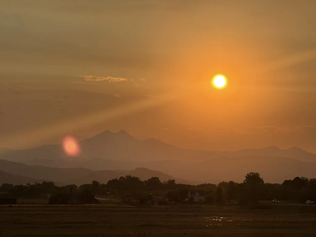 A sun setting over mountains and a field with a dim house in the background.