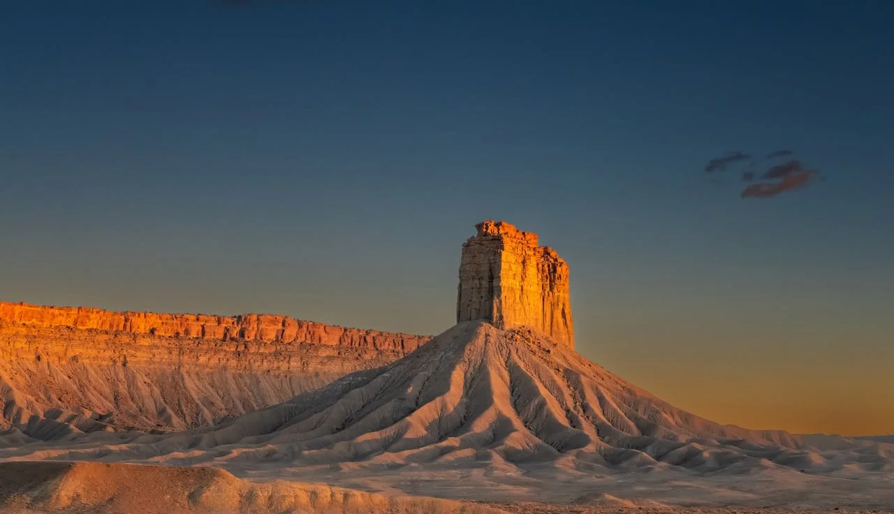 A tall red-toned rock formation surrounded by mesas and a clear blue sky.