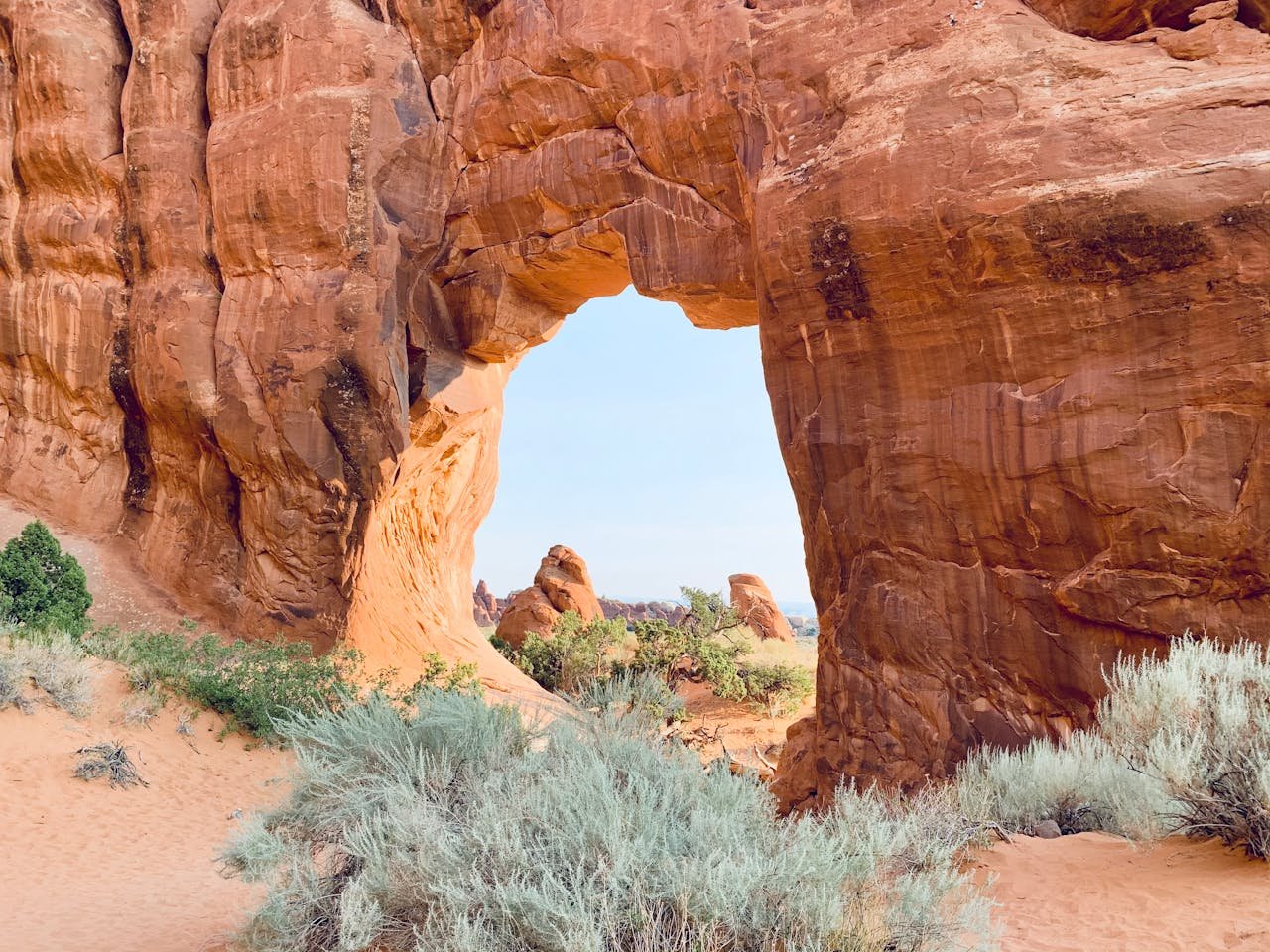 A red-toned rock formation with a large gap inside surrounded by sand and shrubbery