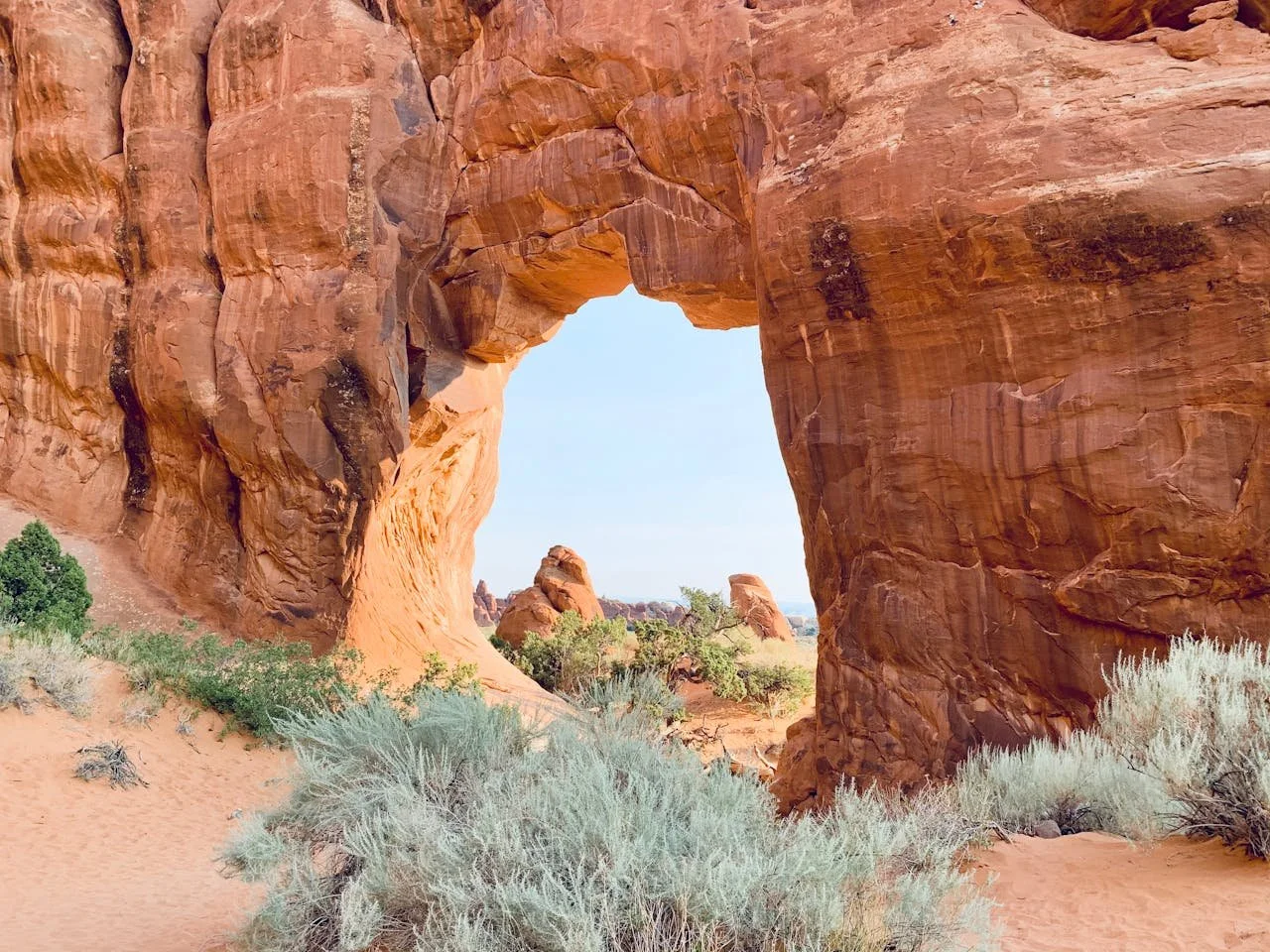 A redrock arch formation surrounded by sand and desert shrubbery.