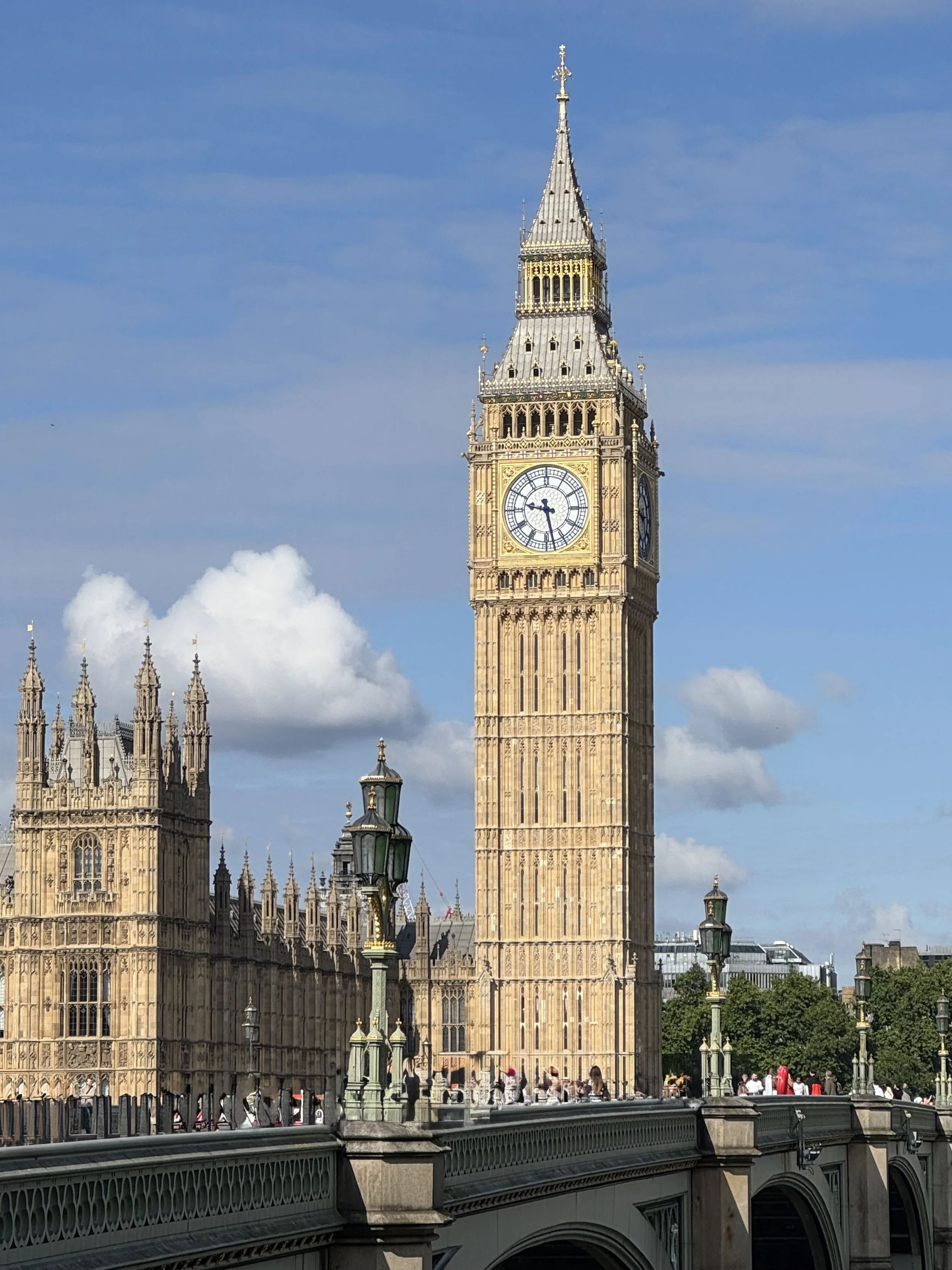 Big Ben clock tower in London with a blue sky and white clouds in the background, with some people walking on the bridge and greenery at the base.