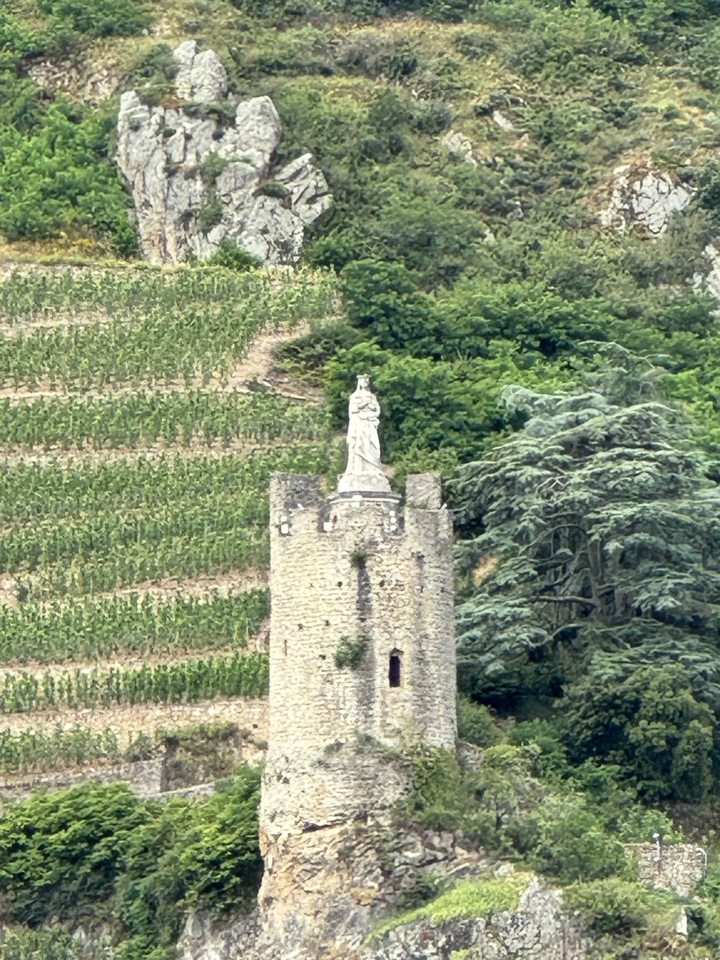Stone tower with a statue on top, surrounded by green trees and vineyard terraces on a hillside.
