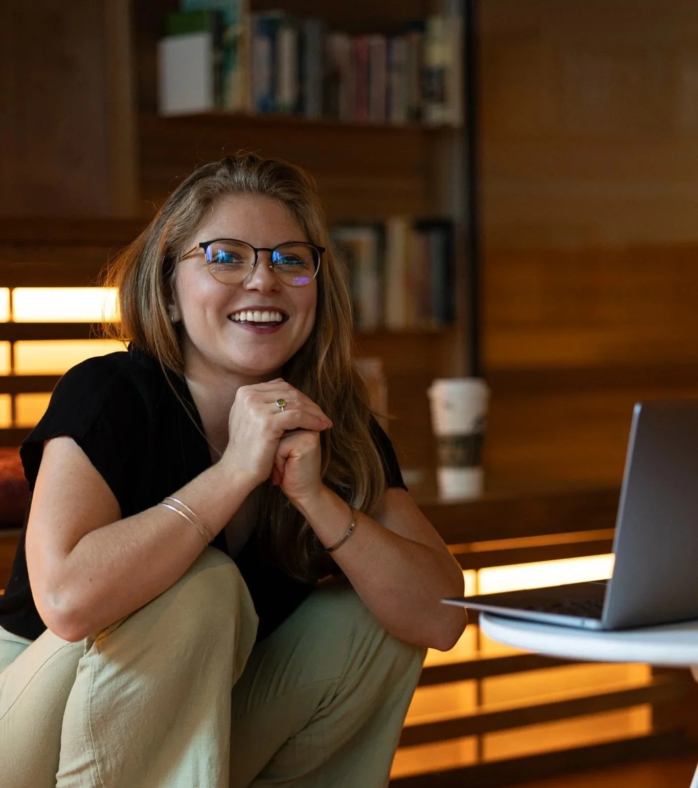 A smiling woman with glasses sitting in a cozy, wood-paneled room with a laptop and a coffee cup in the background.