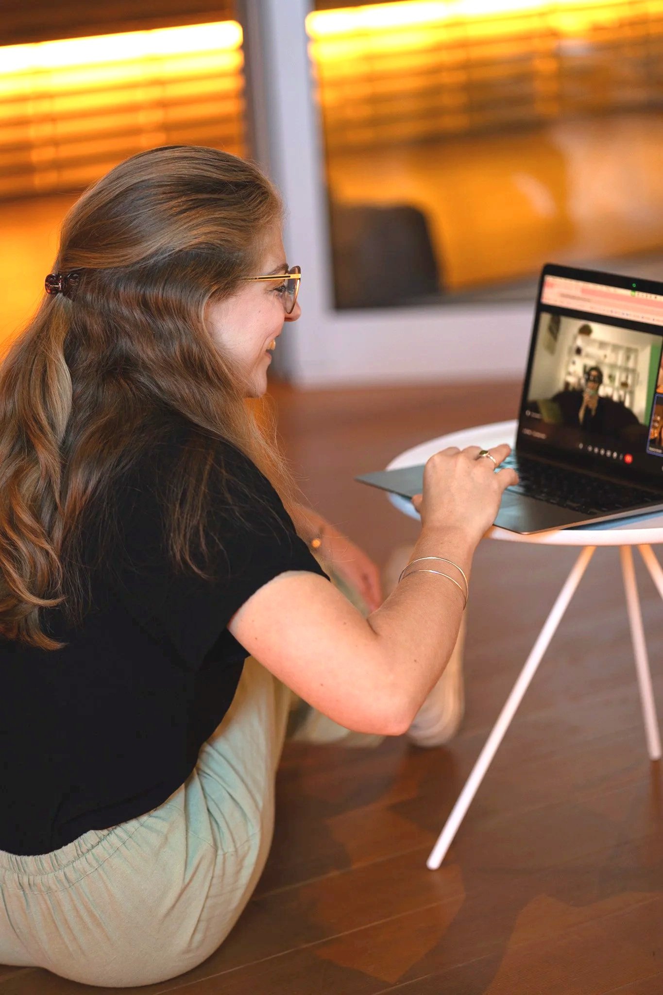 A woman with long hair and glasses sits on the floor, smiling, while participating in a video call on her laptop in a warmly lit room.