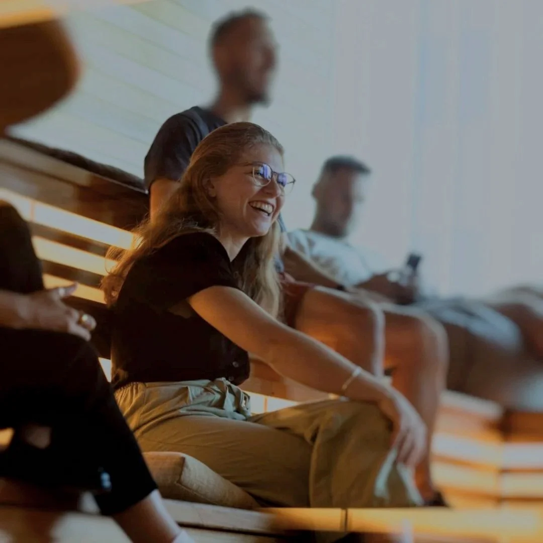 People sitting on benches in a room, smiling and engaging, with a woman with glasses in focus.