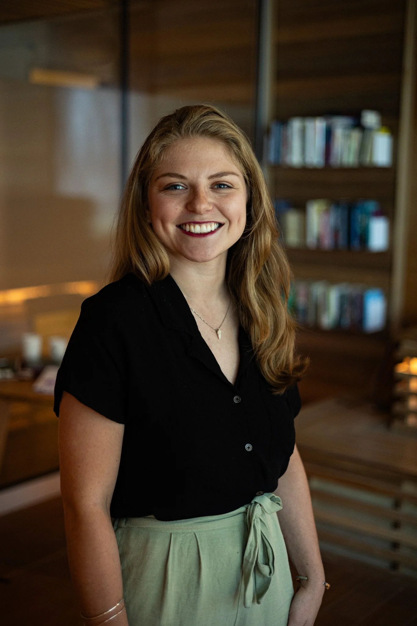 A young woman with long, wavy blonde hair and blue eyes smiling in an indoor setting with wooden walls and bookshelves in the background.
