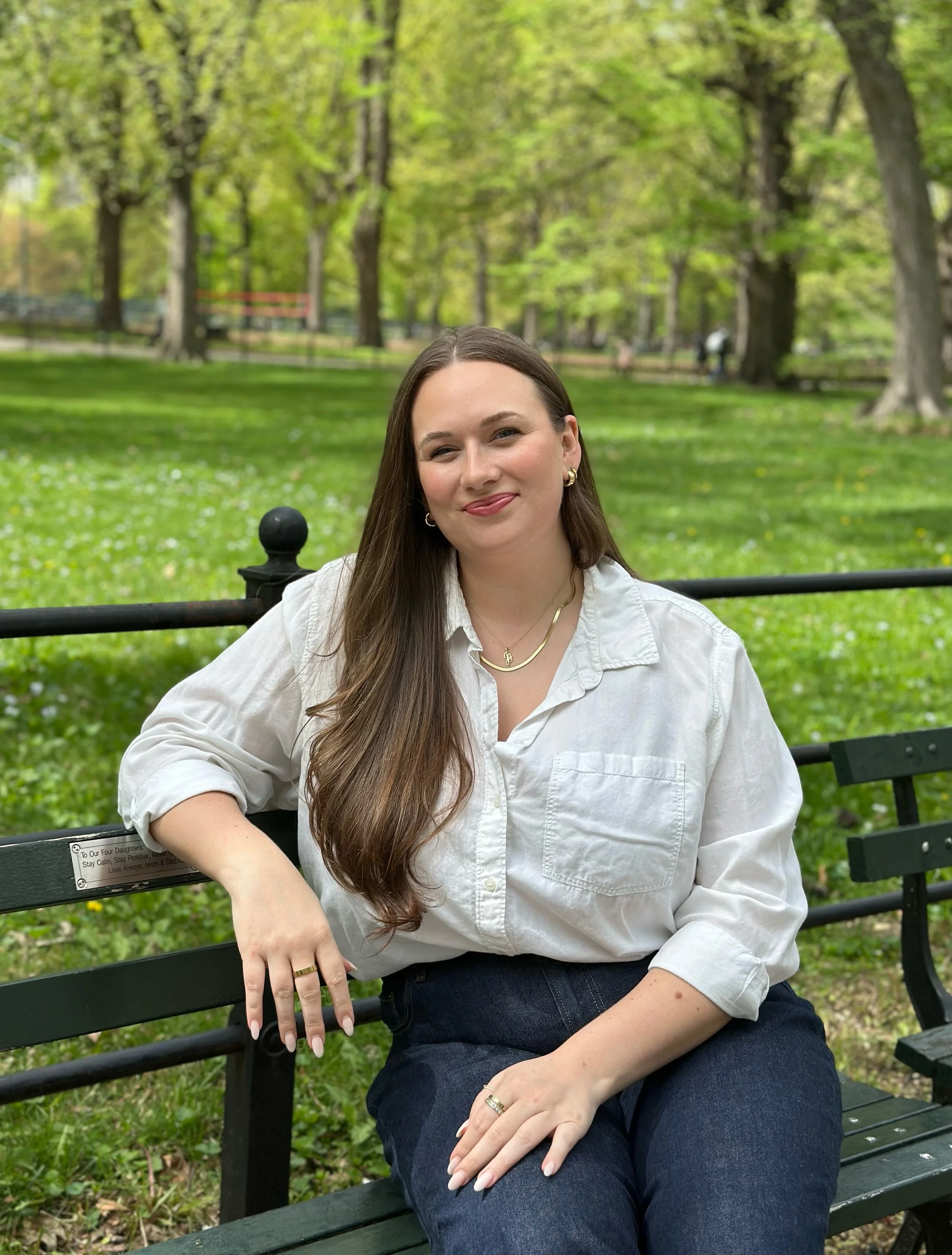 A woman with long hair sitting on a park bench, smiling at the camera, in an outdoor park setting.