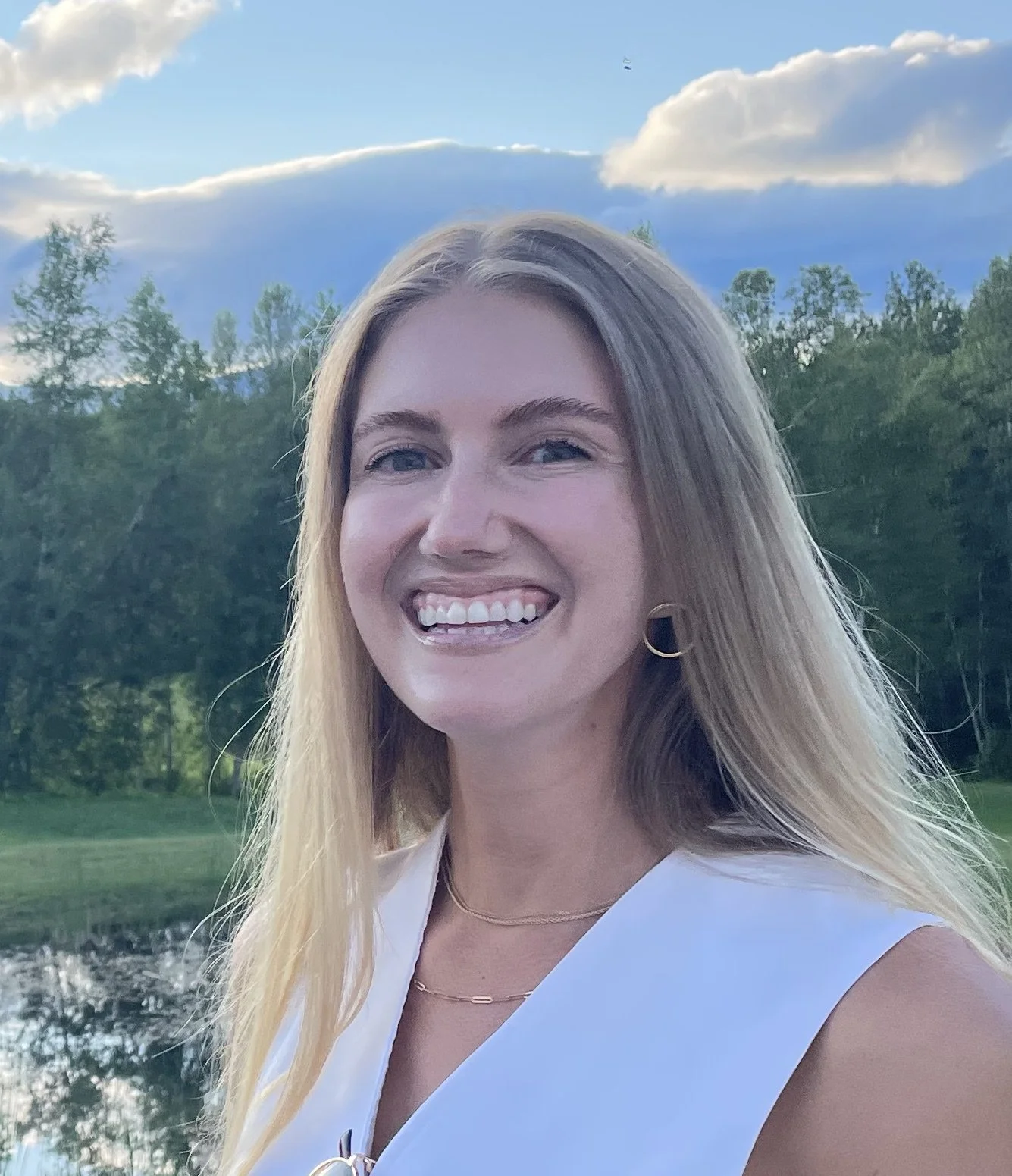 Black and white photo of a smiling woman with long hair, wearing hoop earrings and a sleeveless top, standing outdoors near a body of water with trees and cloudy sky in the background.