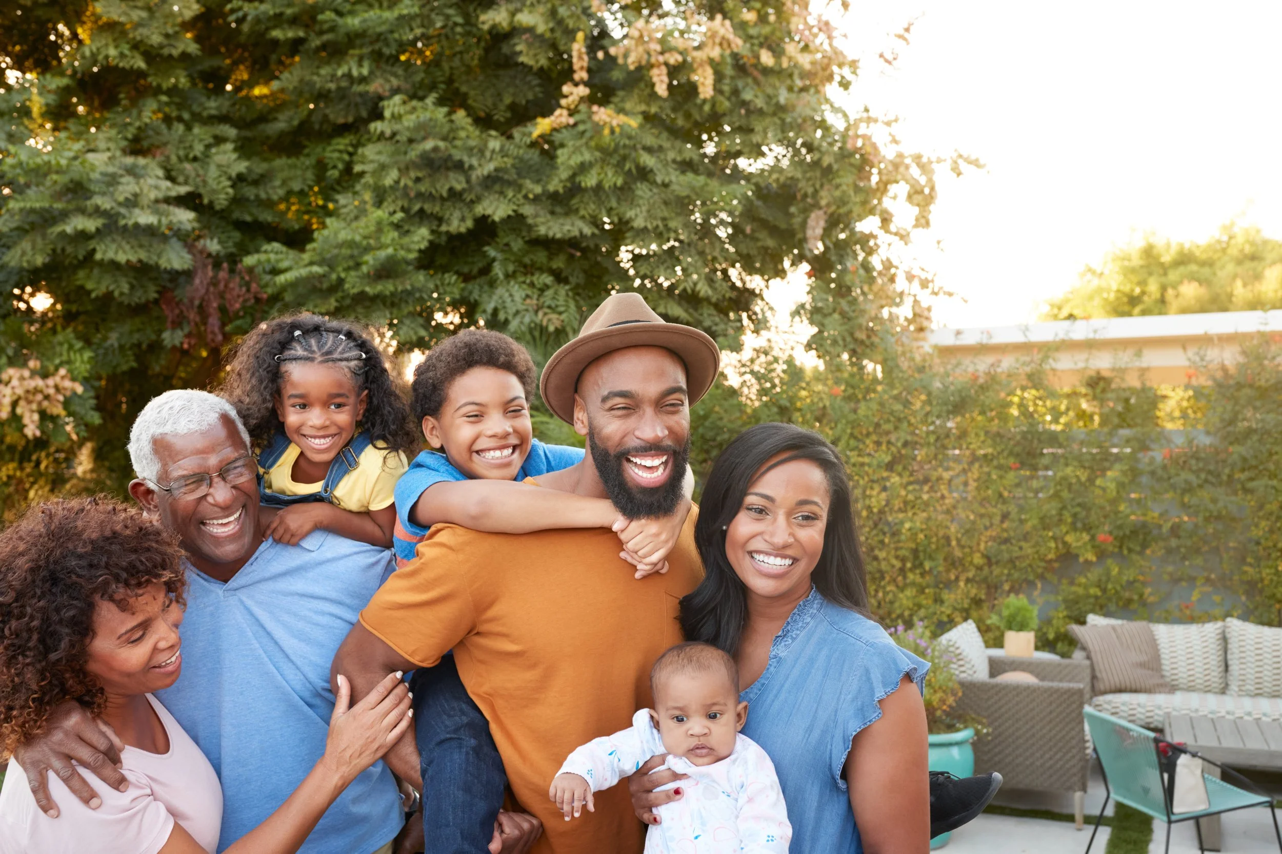 A multi-generational family outdoors smiling, with a man giving a piggyback ride to two kids, others gathered around, in a backyard with trees and outdoor furniture.