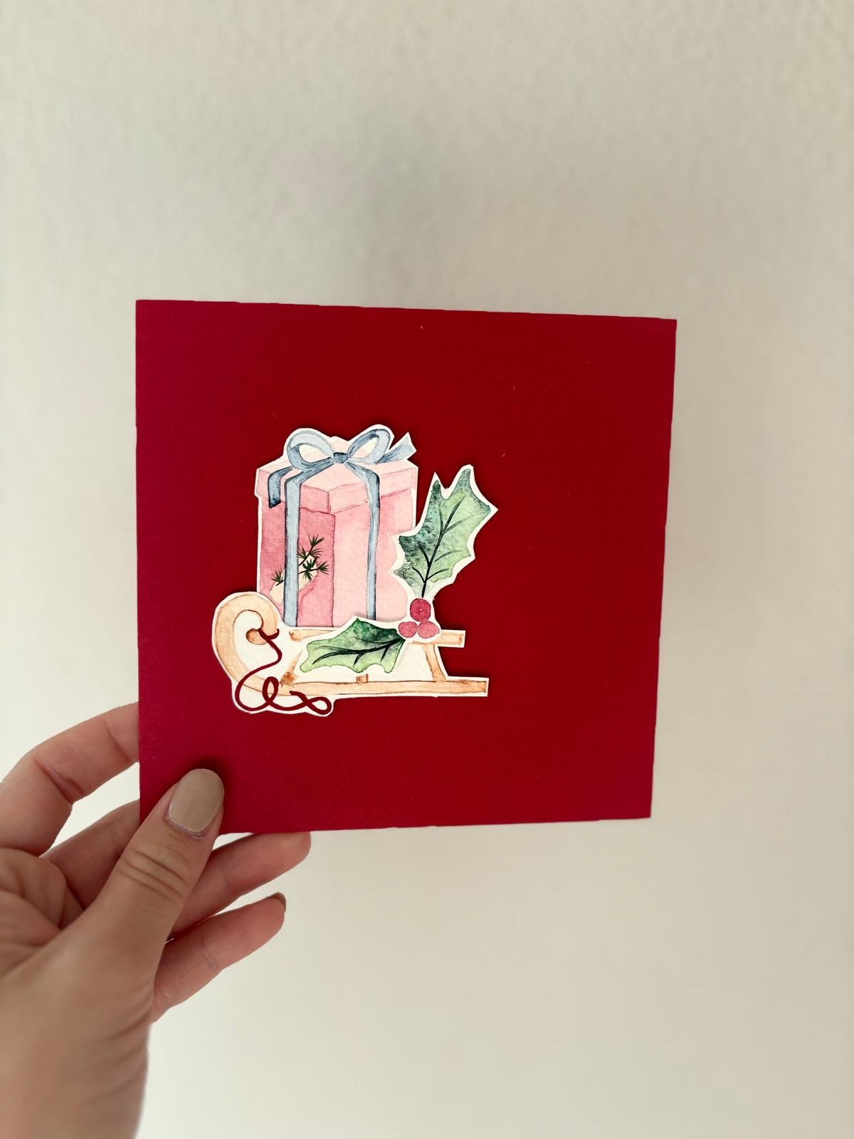 Hand hält eine rote Grusskarte mit einer Illustration aus rosa Geschenk, Holzschlitten und Stechpalmenzweigen Text auf Bild: Love