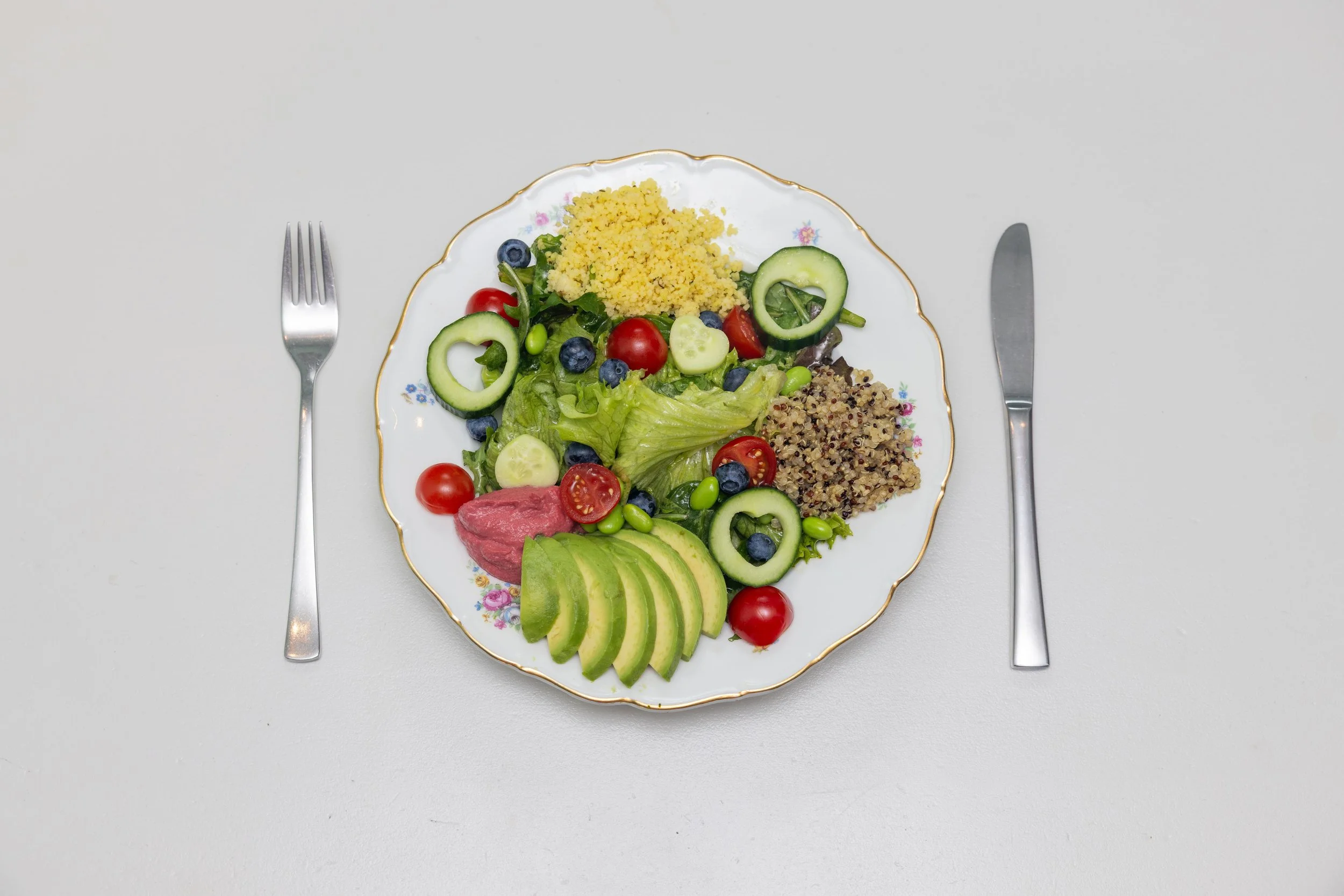 Teller mit Salat, Avocadoscheiben, Tomaten, Gurke, Heidelbeeren, Edamame und Getreidebeilagen, daneben Gabel und Messer auf weissem Tisch
