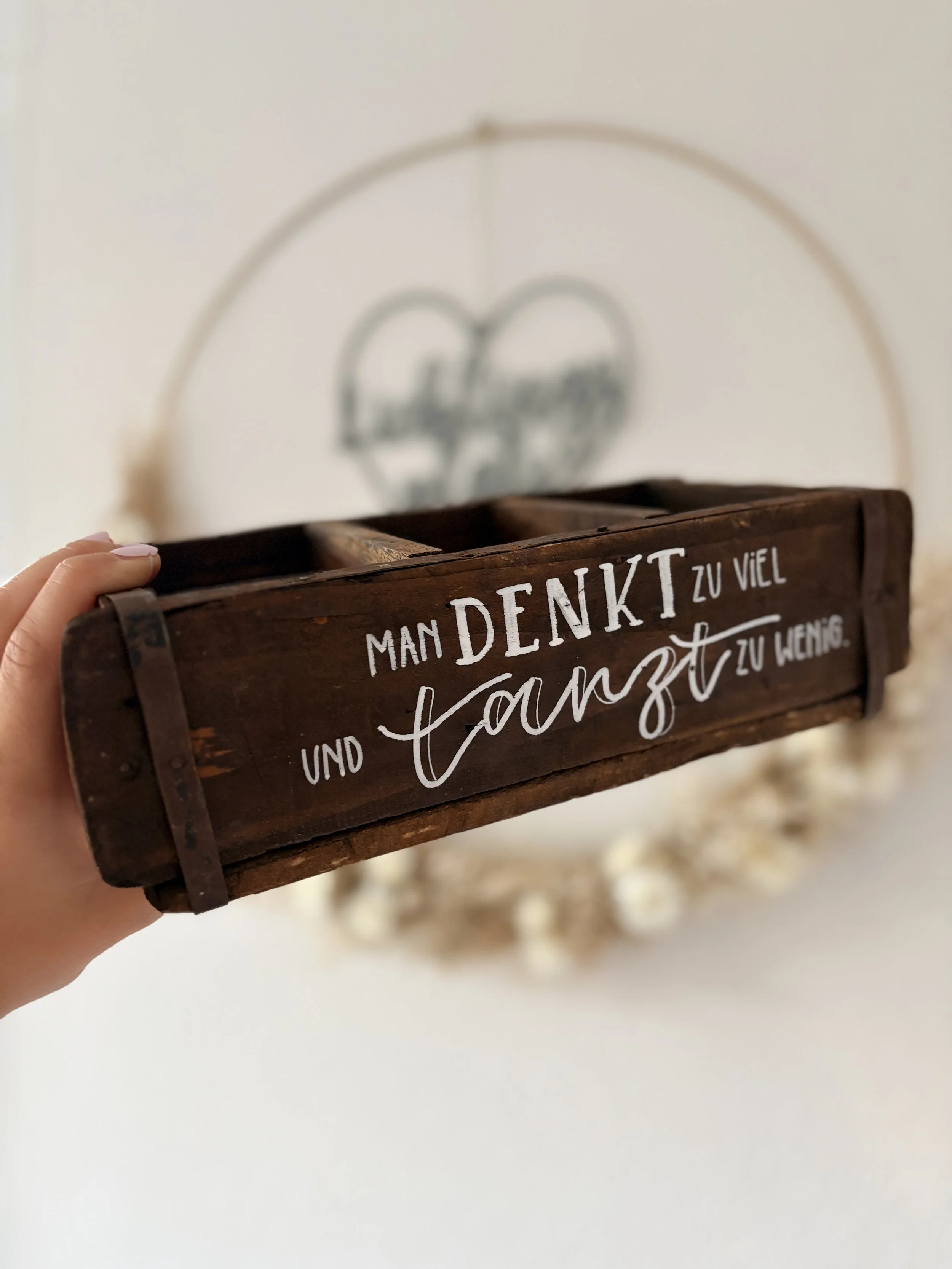 Hand hält dunkle Holzkiste mit weissem Schriftzug, im Hintergrund ein Makramee-Reifen Text auf Bild: Man denkt zu viel und tanzt zu wenig.